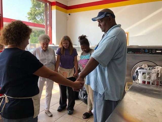A group of five diverse people holding hands in a circle inside a laundromat, with a man and woman at the front.