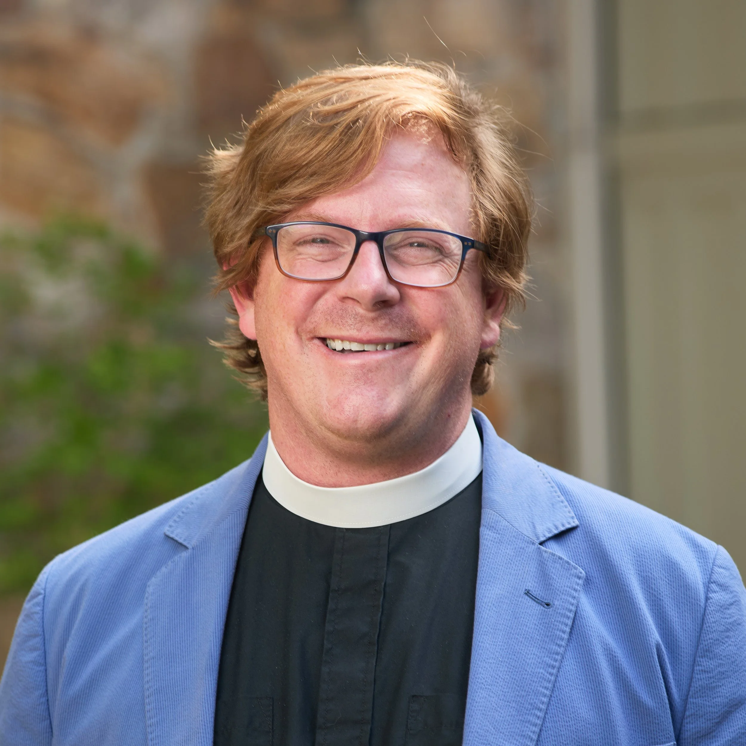 Smiling man in clerical collar and glasses, wearing a blue blazer, outdoors with blurred greenery and building background.