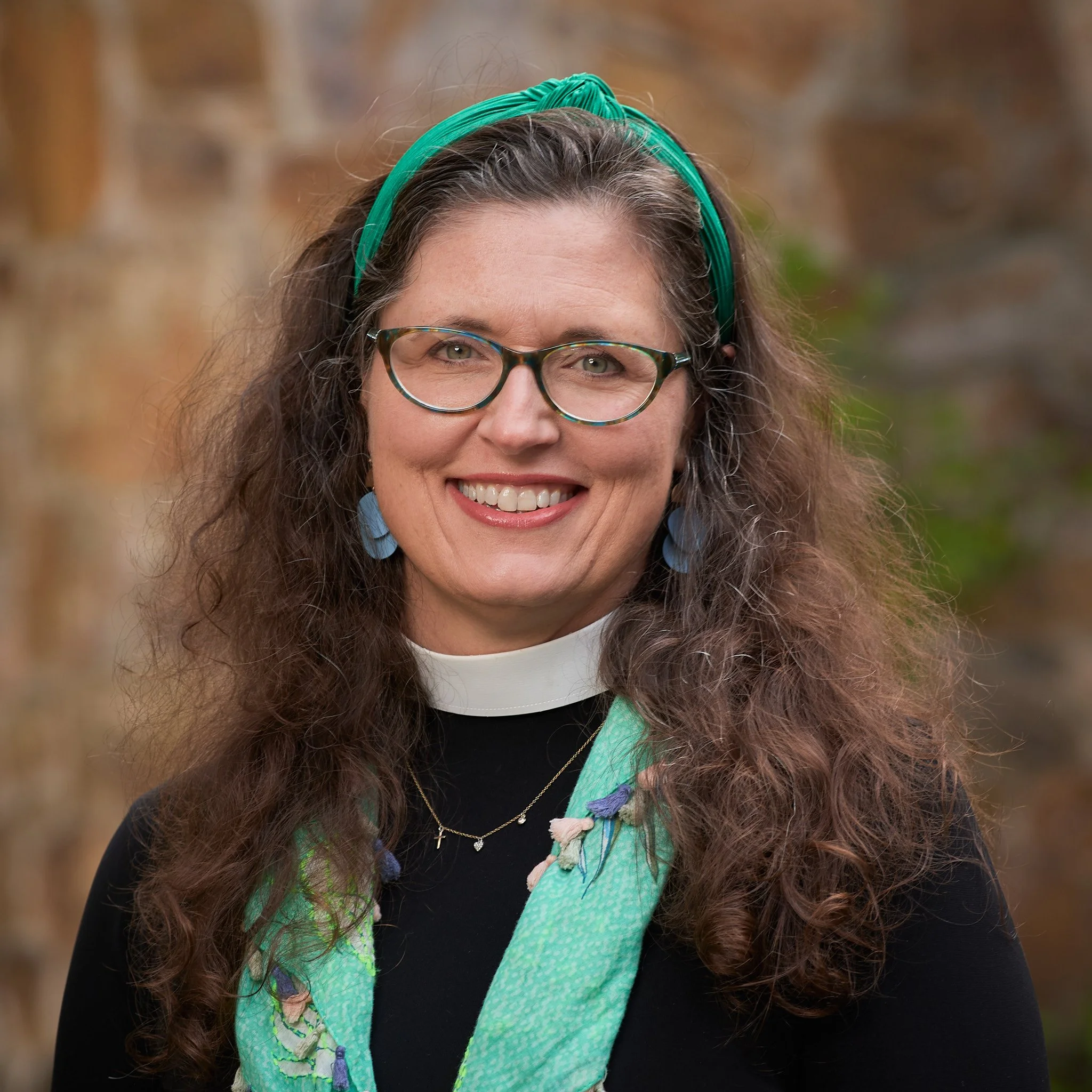 A smiling woman with long, curly brown hair, glasses, and wearing a teal headband, earrings, a black top, a white clerical collar, and a green scarf with purple and pink accents, standing outdoors in front of a blurred stone background.
