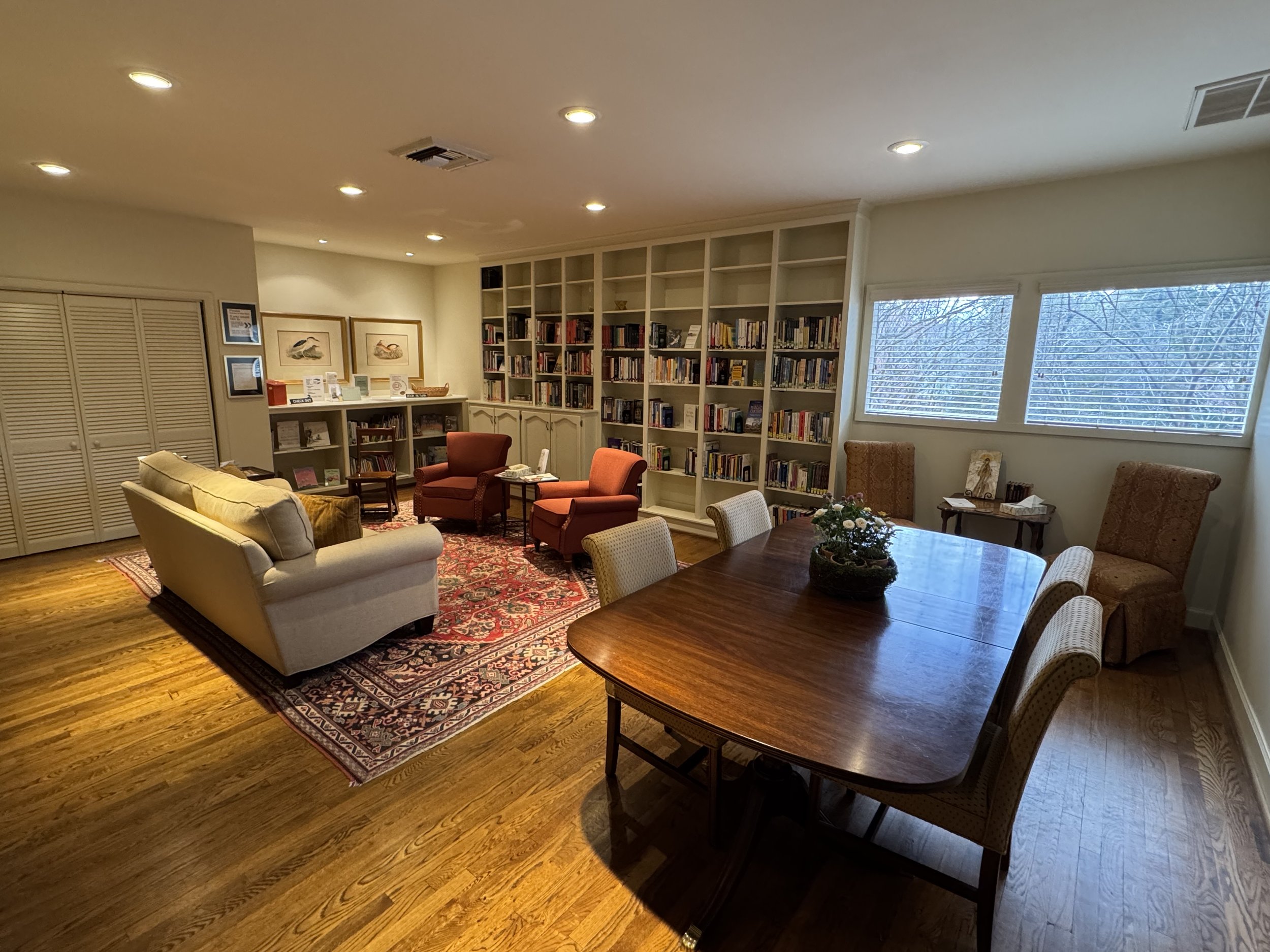 A cozy living room with a beige sofa, two red armchairs, a wooden dining table with six chairs, a bookshelf filled with books, a window with blinds, and artwork on the walls.
