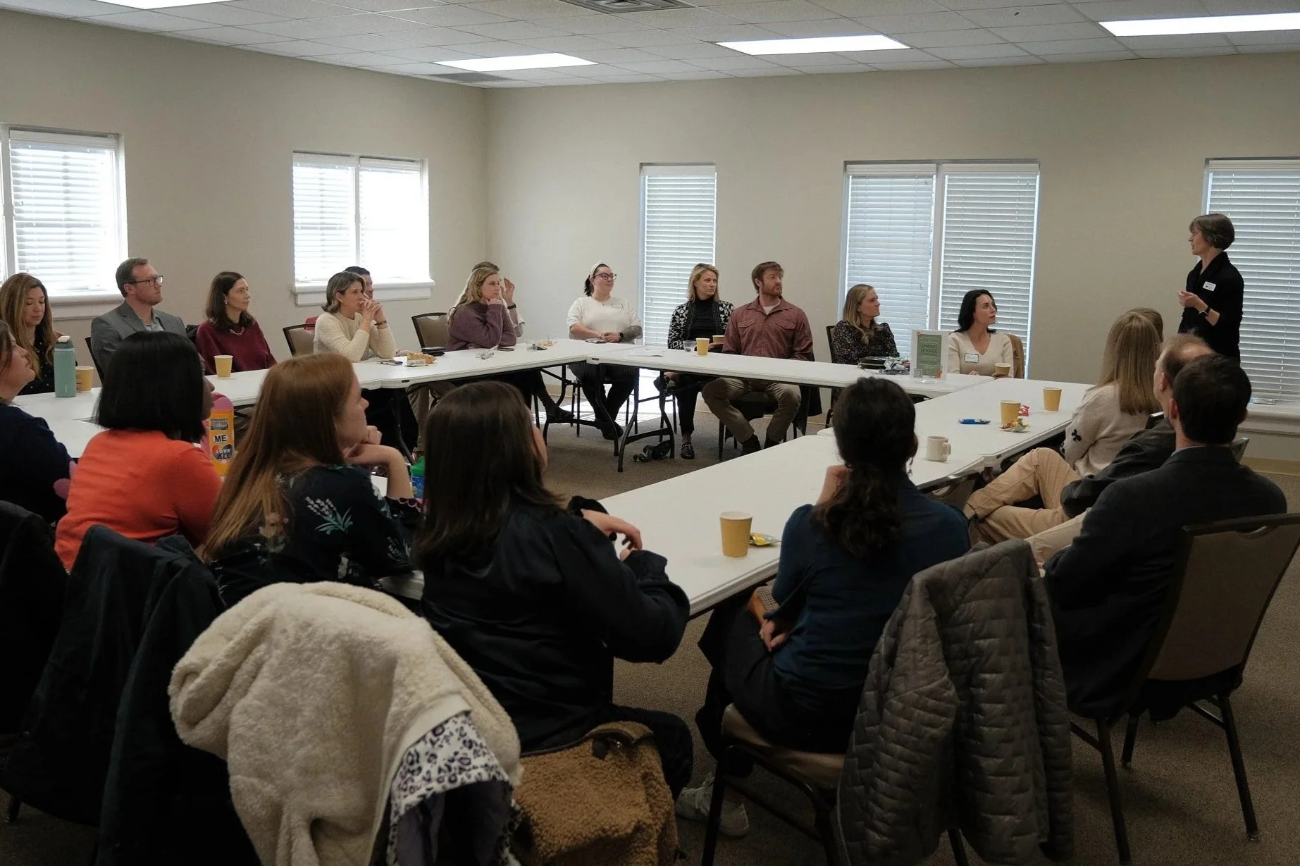 A woman standing and speaking to a group of people seated at U-shaped tables in a conference room with beige walls and five windows with blinds.