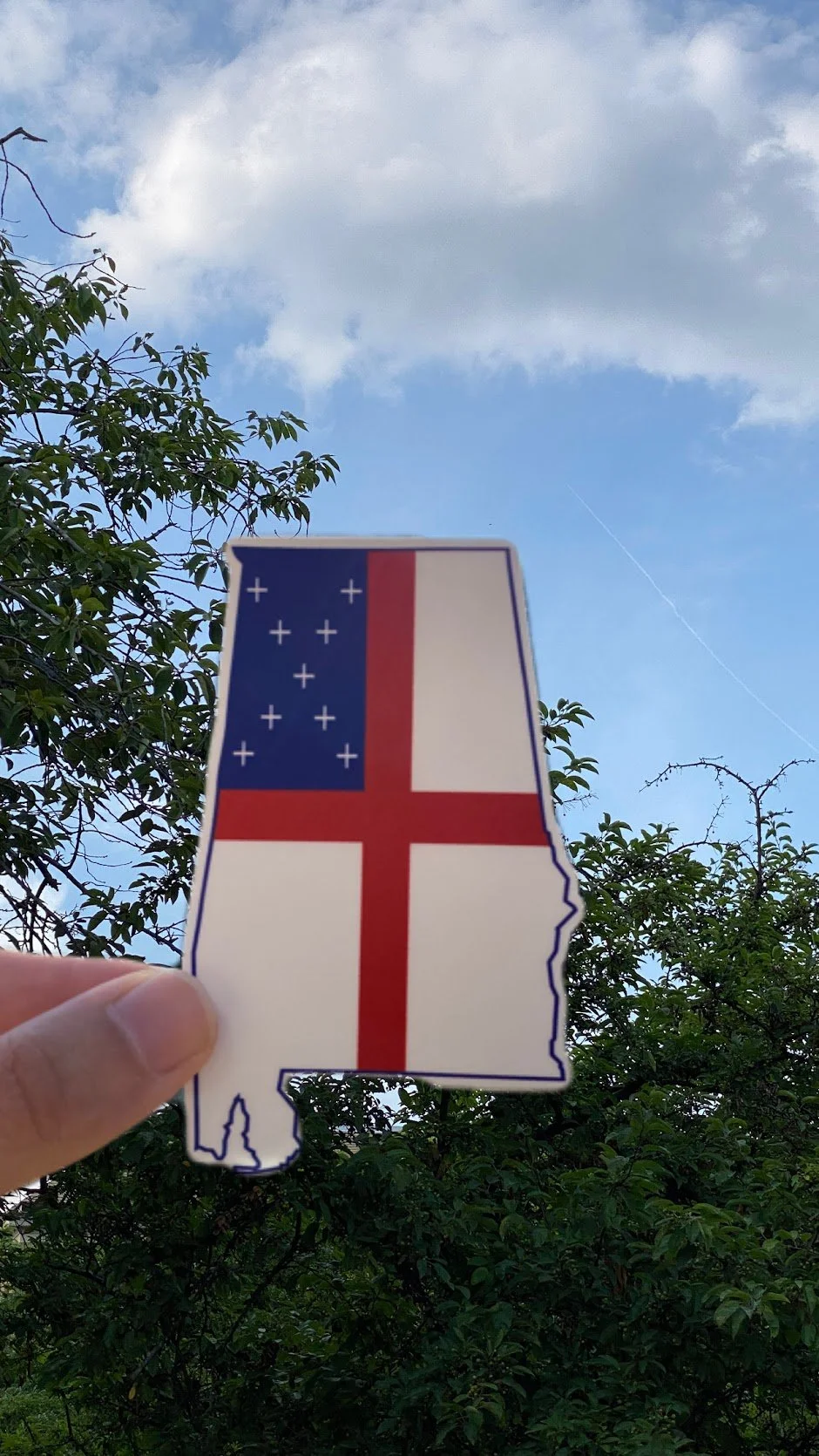 Hand holding a sticker in the shape of the state of Connecticut with the design of the American flag against a backdrop of blue sky and green trees.