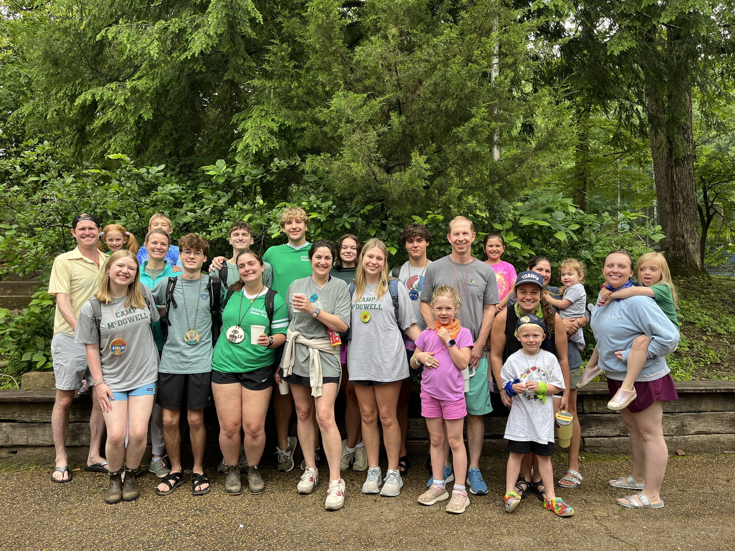 Group of children and adults outdoors in a forested area, posing for a photo with some holding cups and wearing camp T-shirts and leis.