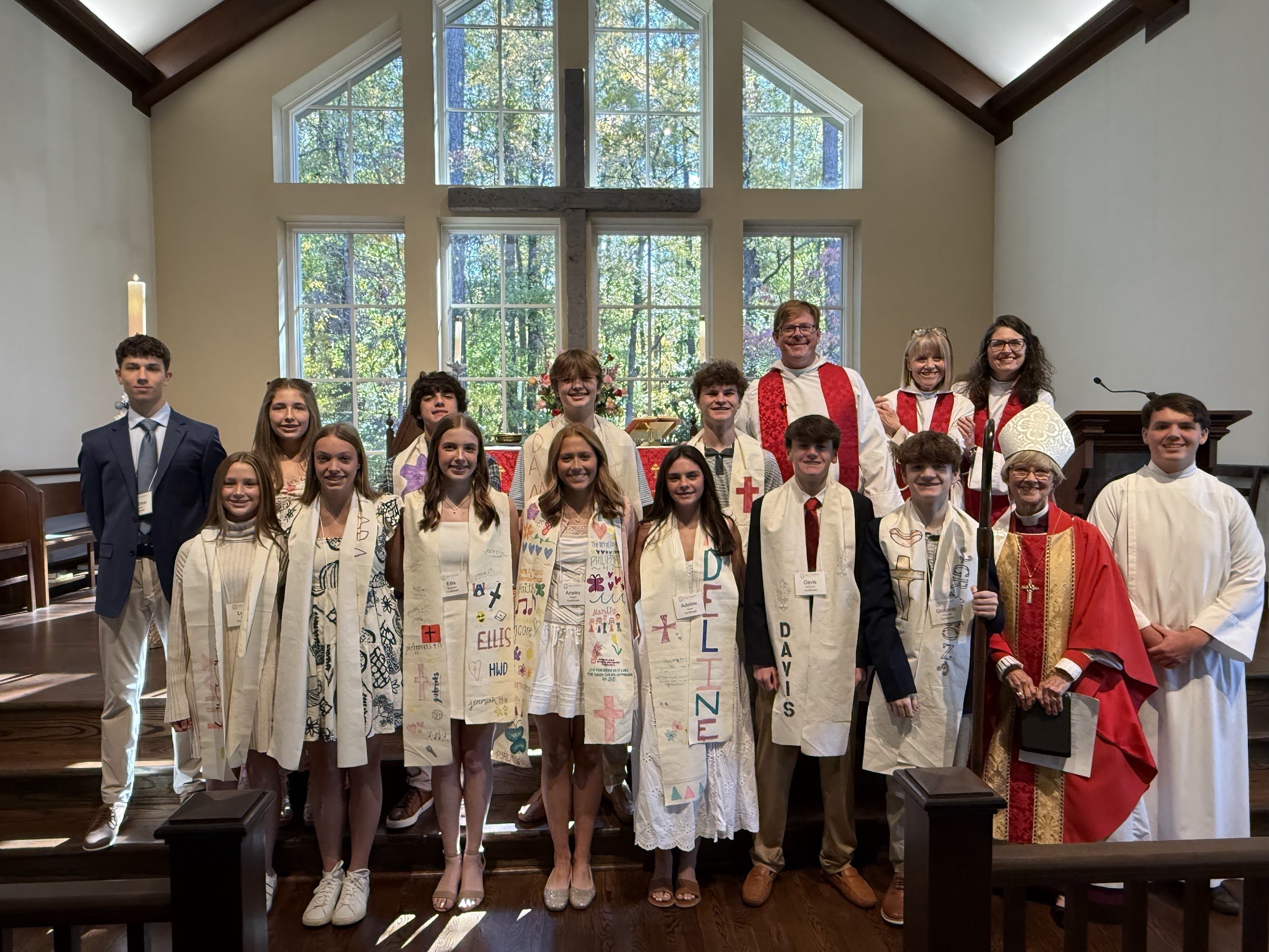 Group of children and clergy members inside a church, some wearing white stoles with names and drawings, standing on steps in front of a large window with trees outside, celebrating a religious ceremony.