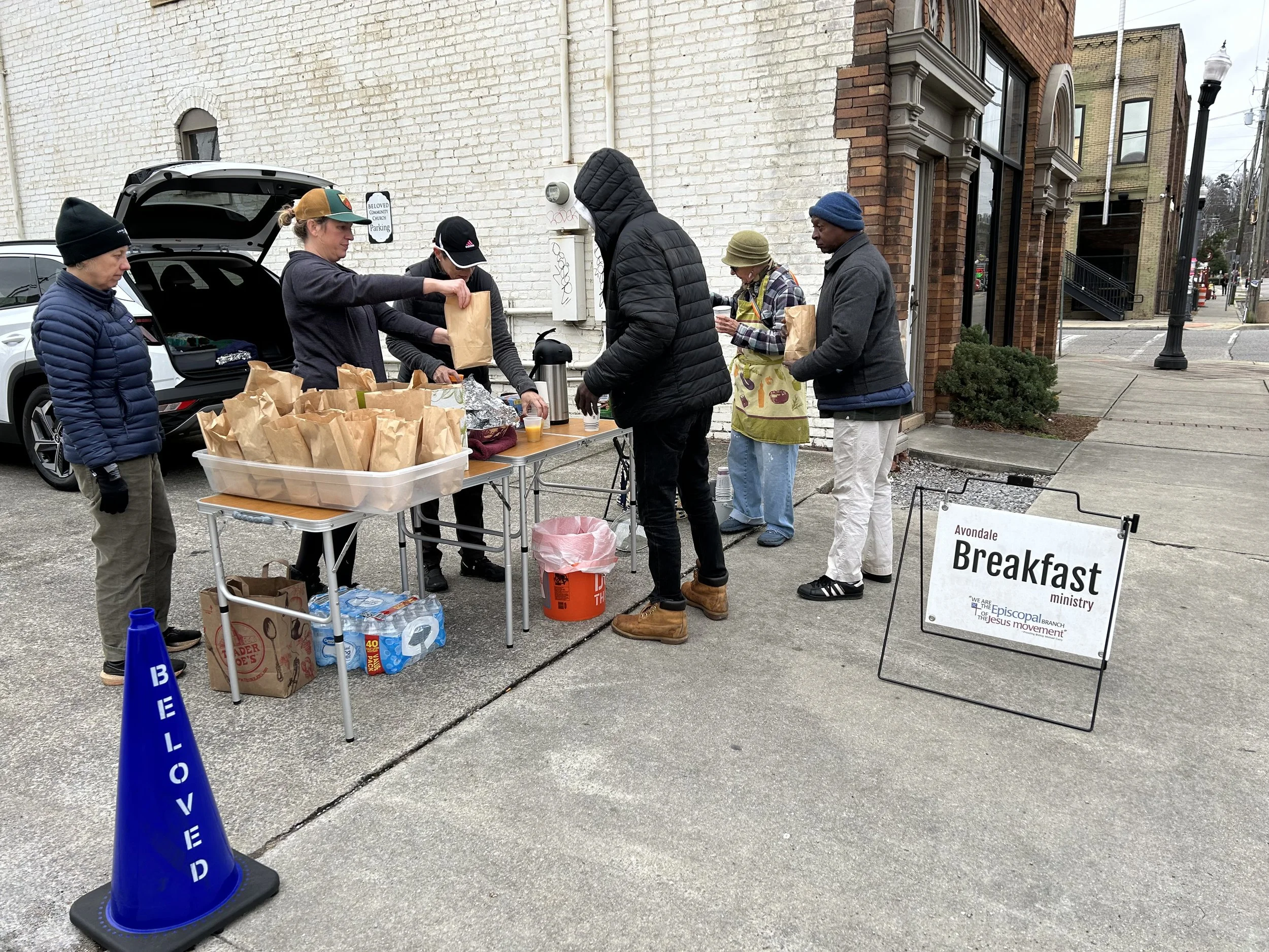 People lining up outside for a free breakfast ministry, with tables holding paper bags of food and a sign reading 'Avondale Breakfast ministry,' set against a brick and white wall in an urban setting.