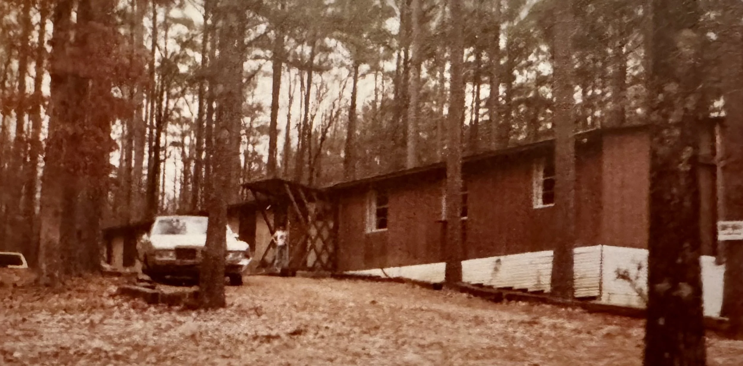 A brick building in a forested area with tall trees and several vehicles parked nearby, including a white van and a truck.