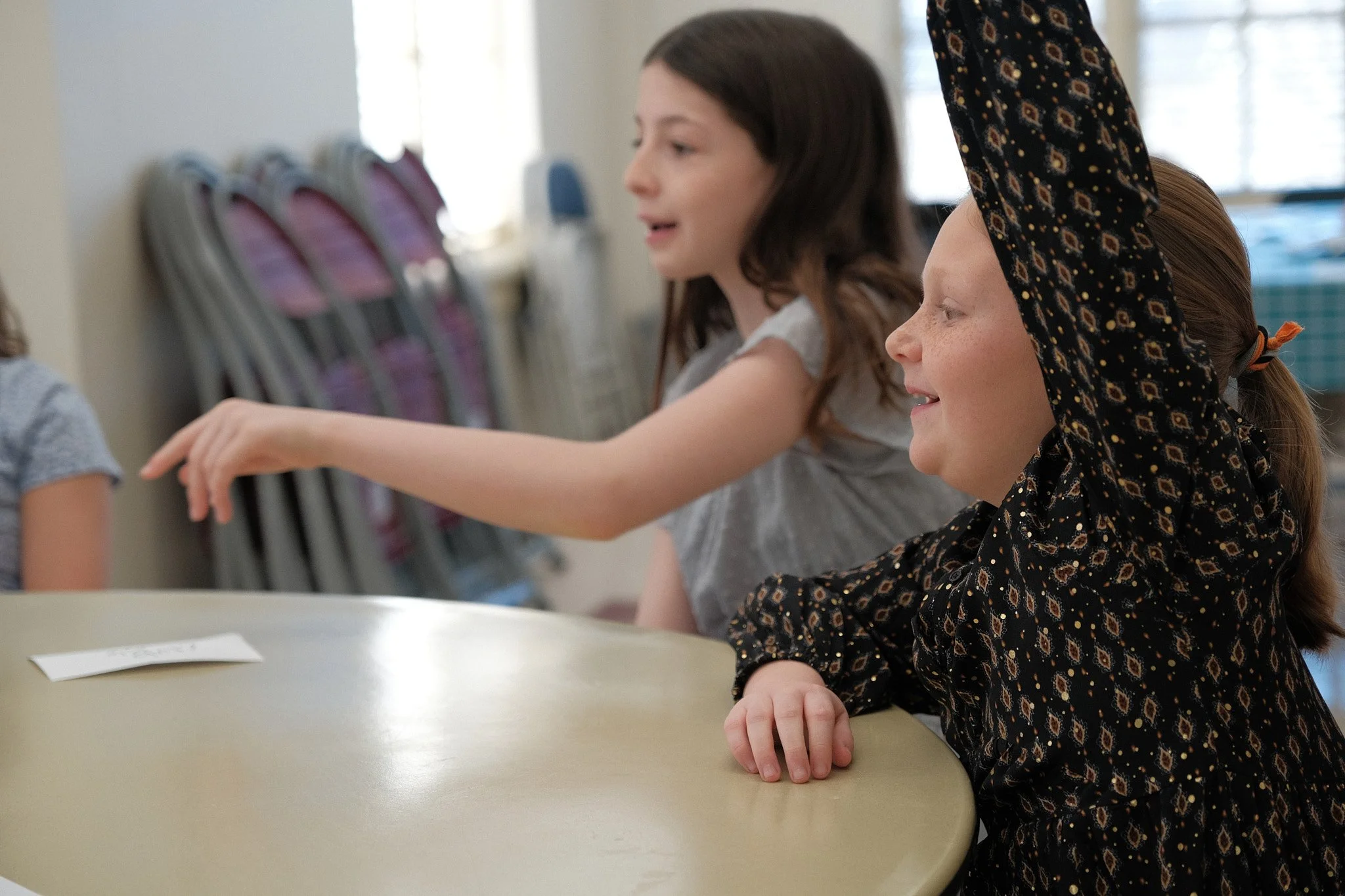 Two young girls sitting at a table, one in a gray shirt with brown hair, the other in a black patterned shirt with a matching headscarf, smiling as they engage in an activity in a well-lit room.