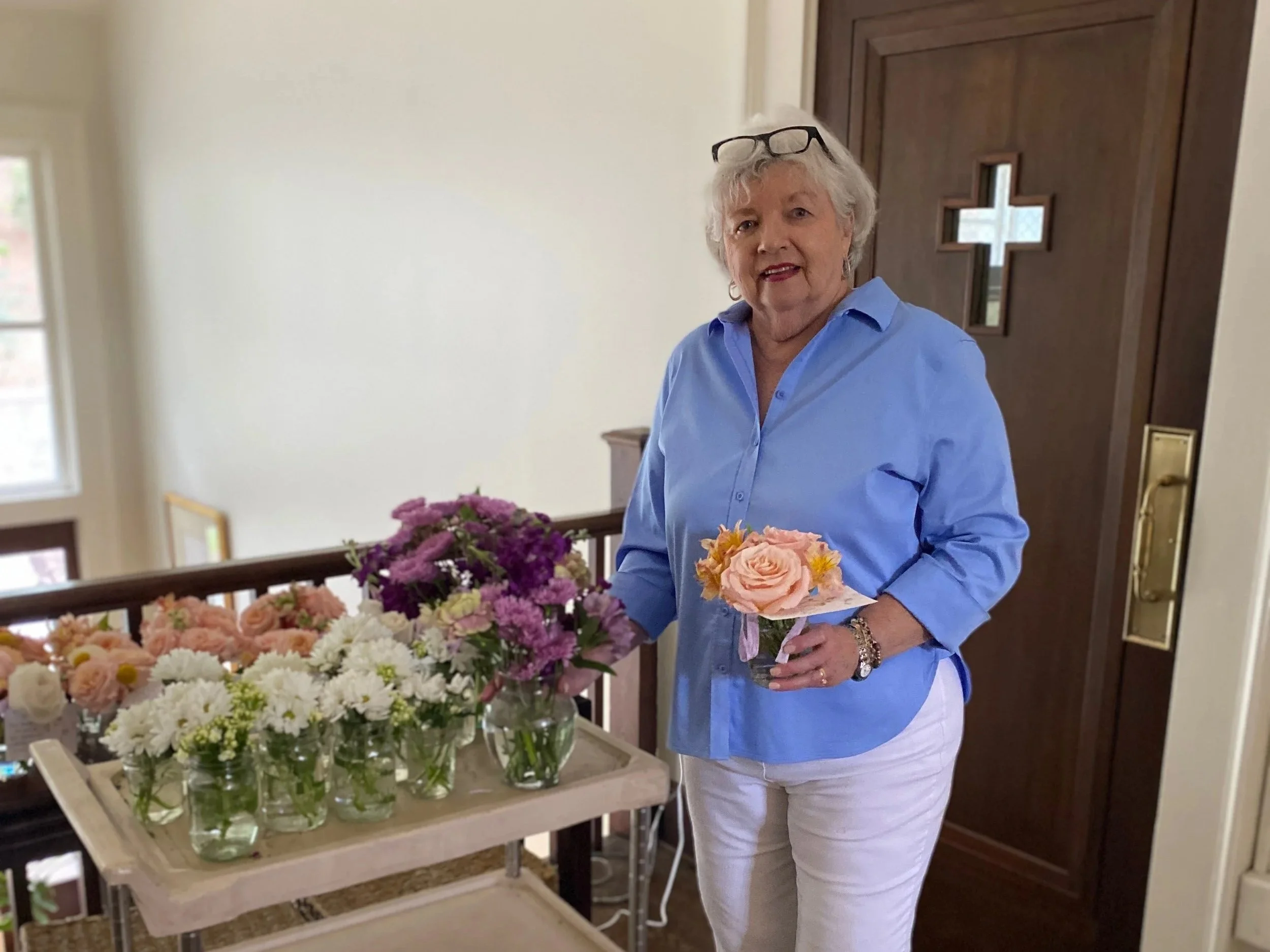 Older woman holding a small bouquet of flowers, standing next to a table with multiple glass vases of colorful flowers, in a well-lit home.