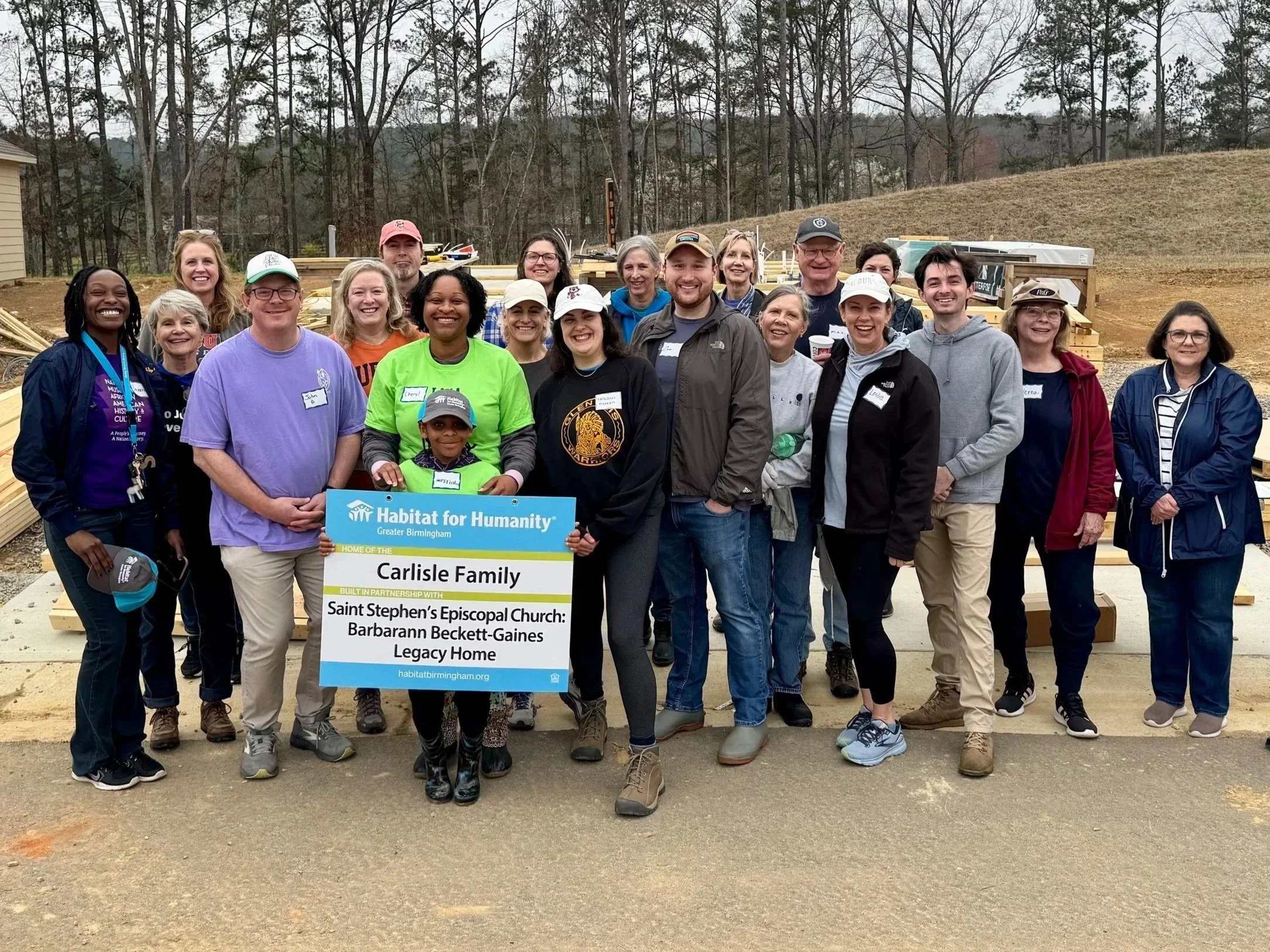 A group of people holding a Habitat for Humanity sign during a building project outdoors, with construction materials and a partially built structure in the background.