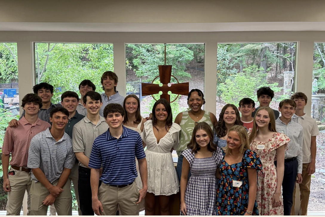 Group of young students and an adult posing indoors in front of a window with a cross and lush greenery outside.