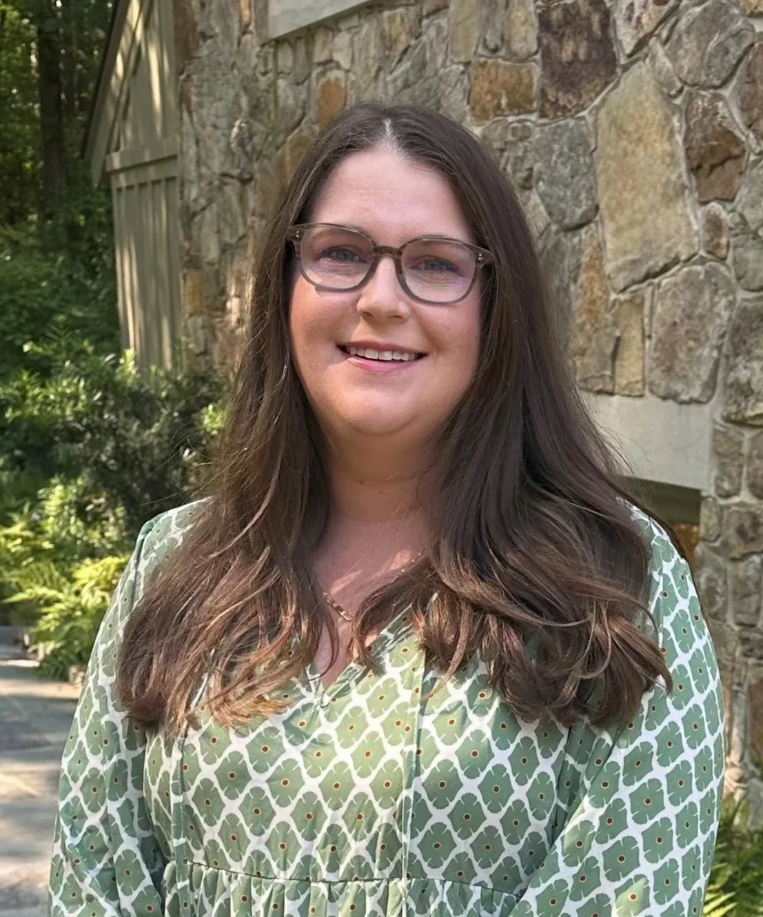 A woman with long brown hair and glasses smiling outdoors. She is wearing a green patterned dress, standing in front of a stone wall and greenery.