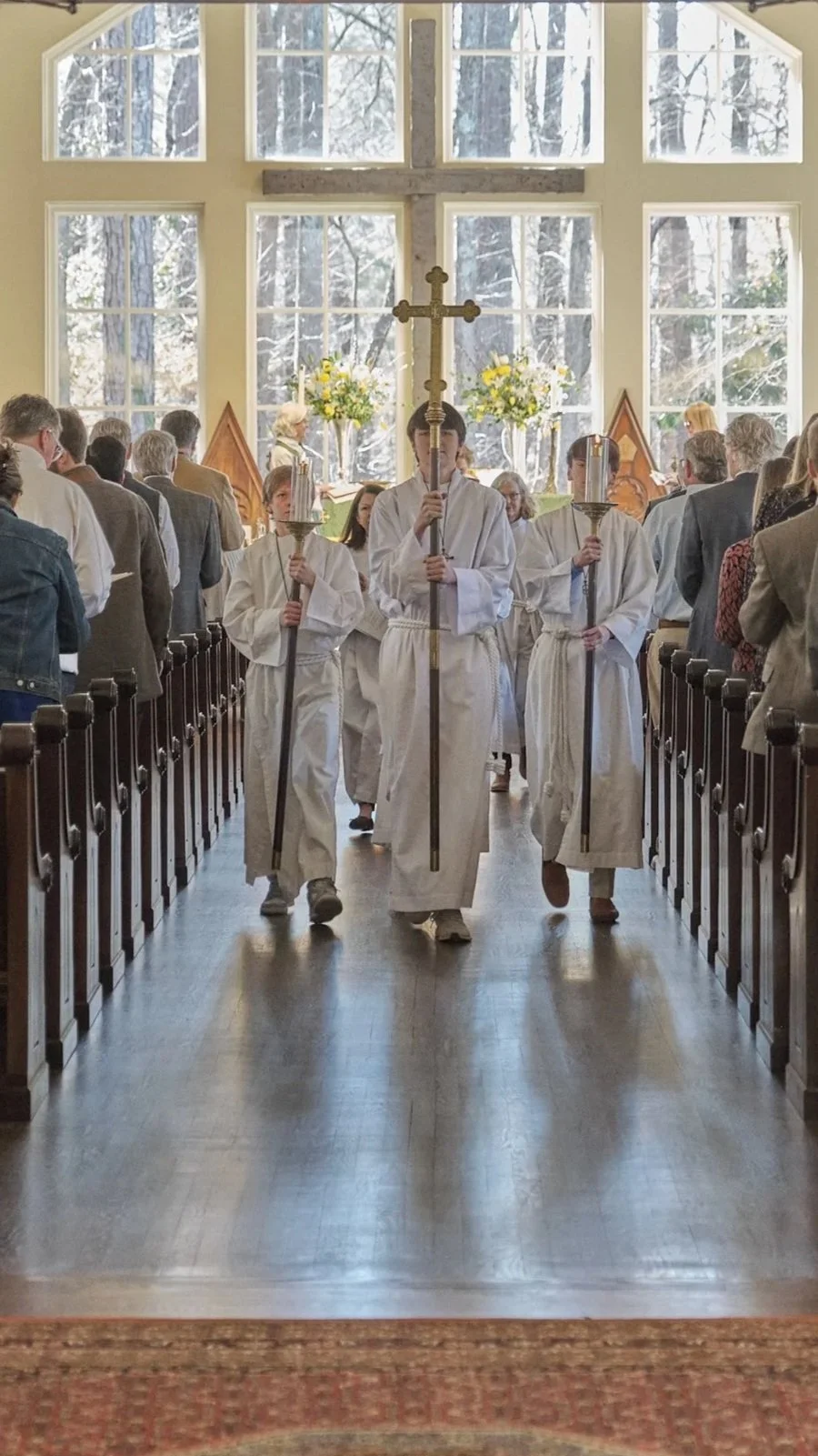Clergy members walking down the aisle of a church during a religious procession, with congregation seated on both sides and large windows showing trees outside.