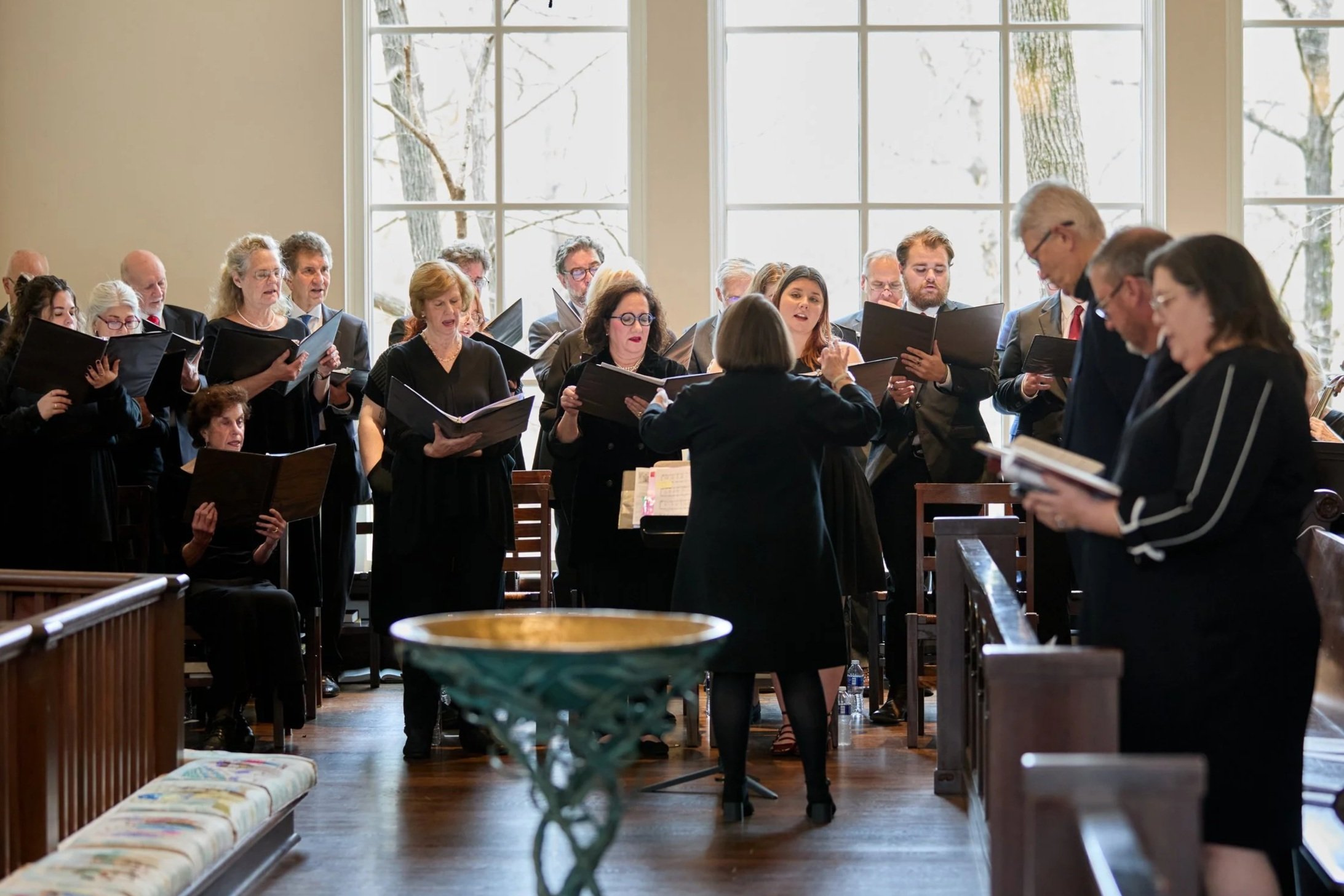 A choir group of men and women singing in a church with a conductor leading them. The choir members hold black folders and are dressed in formal black attire. Large windows behind them let in natural light. Some members are standing while others are seated.
