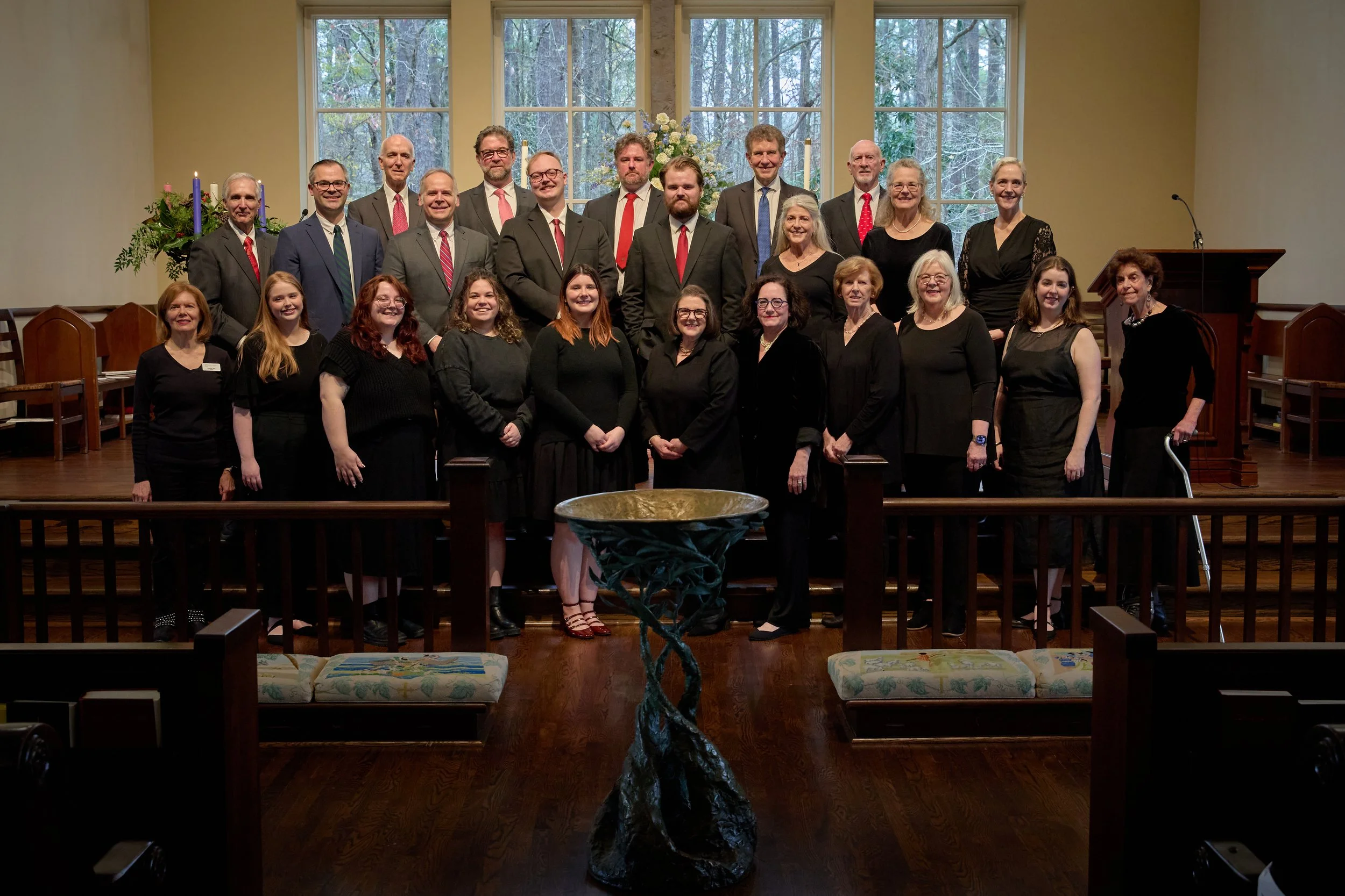 Group of adults standing on a wooden stage in a church or chapel, with large windows showing trees outside. They are dressed in formal or semi-formal black and dark clothing, with some men in suits and ties, women in dresses. A flower arrangement is behind them, and a large baptismal font is in the foreground.