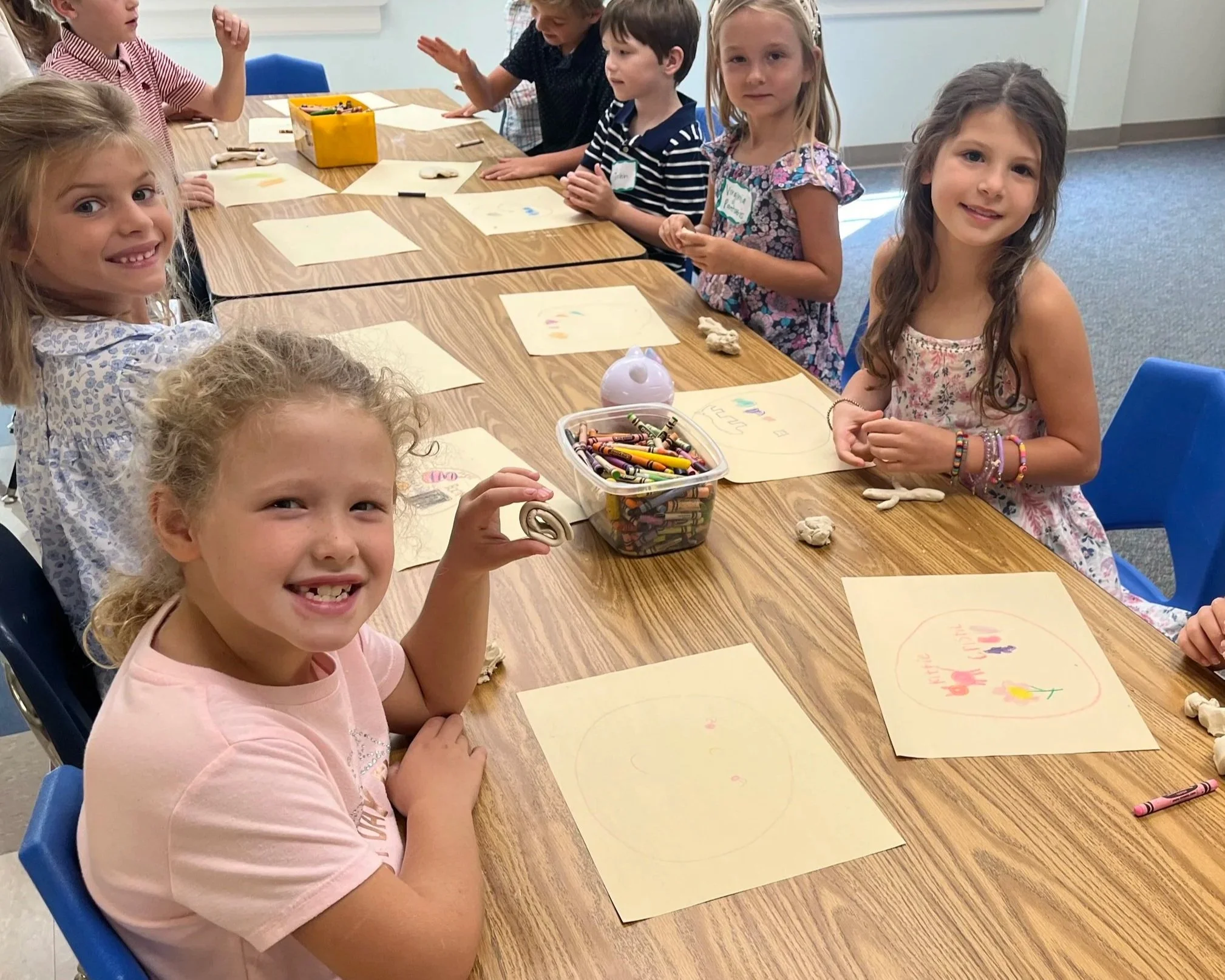 Children sitting at a table with art supplies, drawing and coloring on paper during a classroom activity.