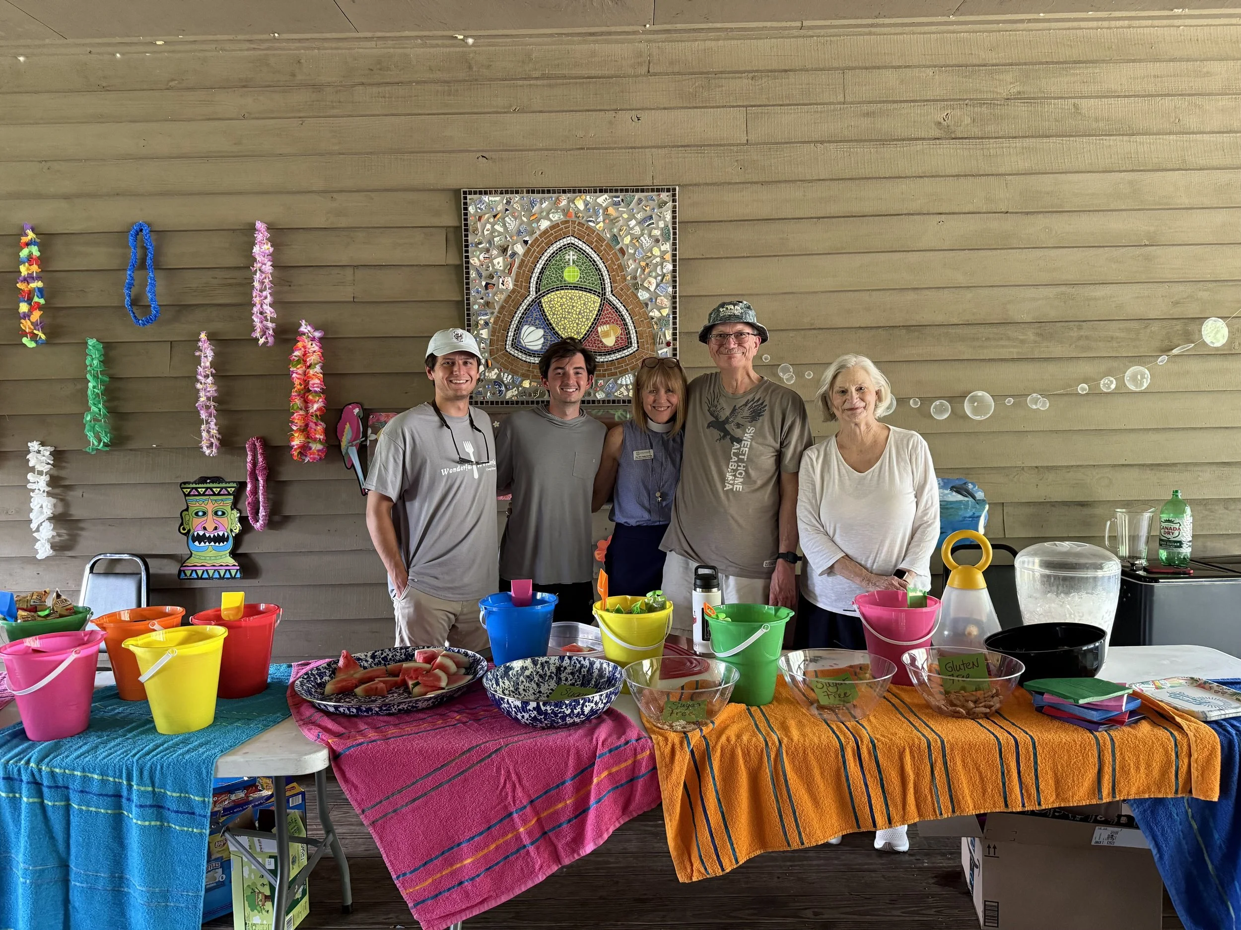 Group of five people standing behind a table with colorful buckets and bowls, tropical drinks, and watermelon slices, decorated with leis, paper decorations, and a mosaic artwork on a wooden wall.