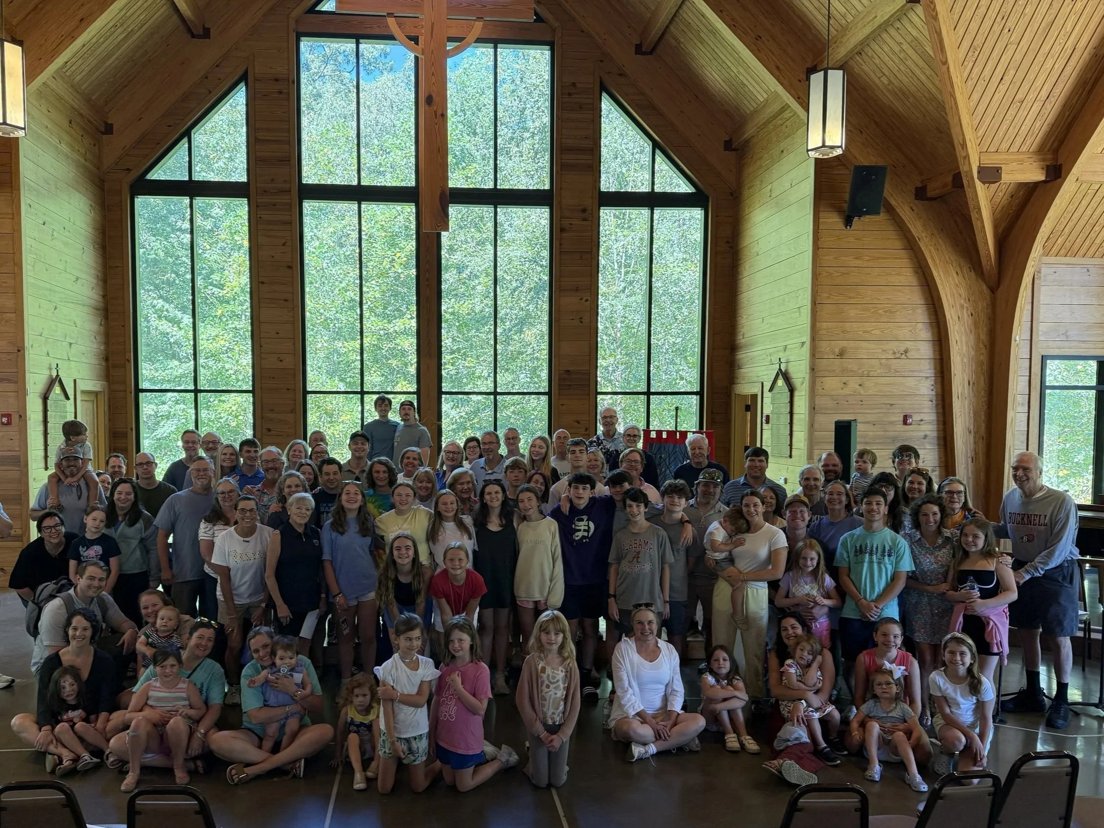 Group photo of a large crowd of people, including children, teenagers, adults, and seniors, inside a wooden building with large windows and trees visible outside.
