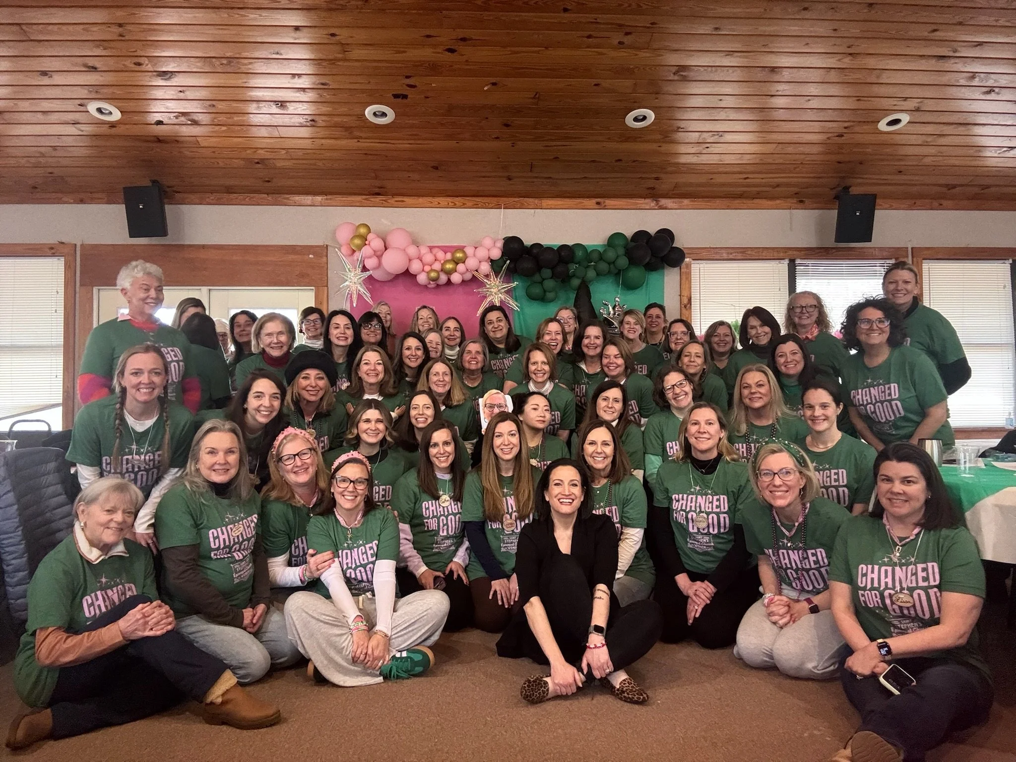 Group of women gathered indoors for a celebration, many wearing matching green shirts with pink and white text, with pink and black balloons and decorations in the background.