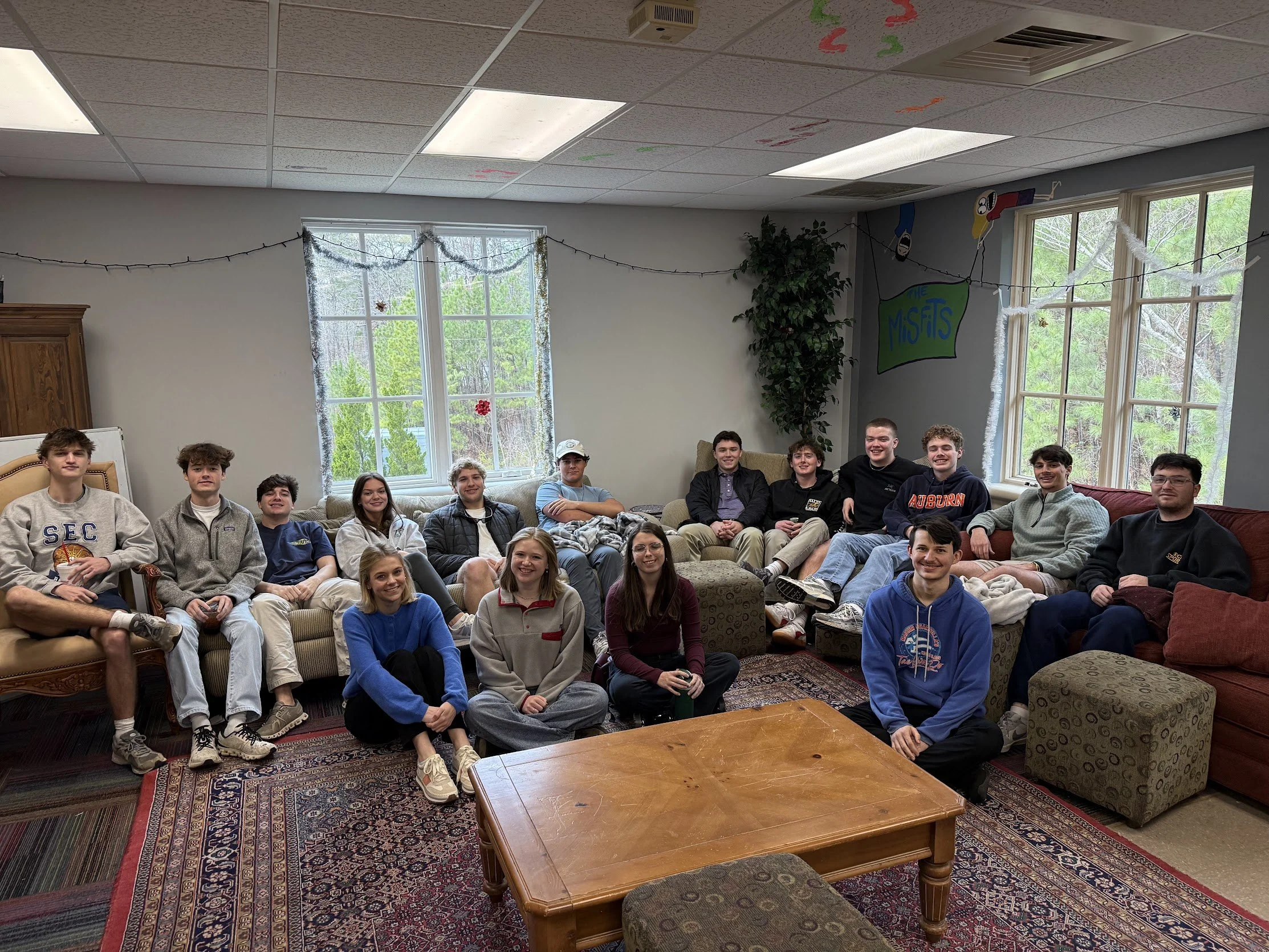 Group of fourteen teenagers sitting and kneeling on sofas and the floor in a living room, with large windows, a carpet, and hanging decorations, including a sign that says 'The Misfits'.