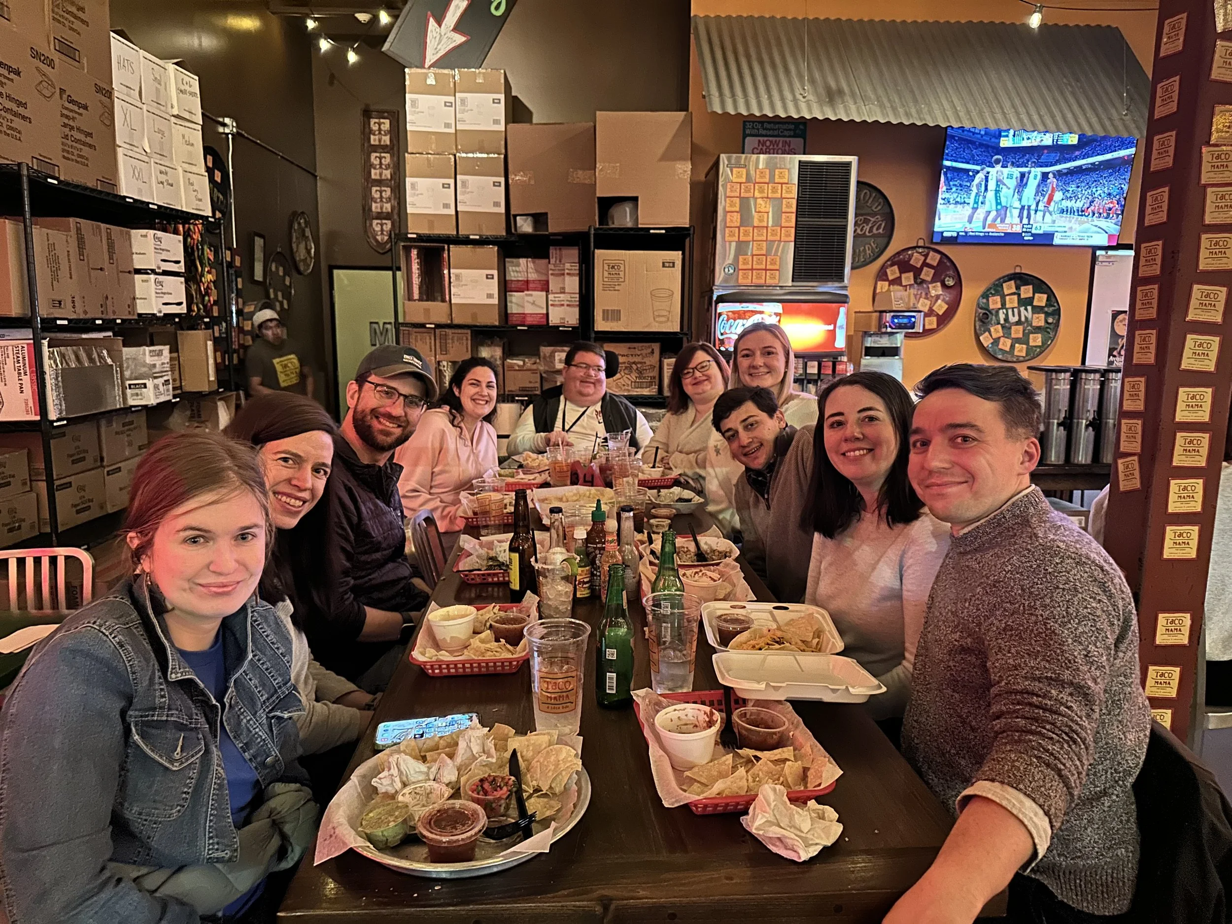 A group of nine people sitting around a table at a restaurant enjoying food and drinks, with a TV showing a sports game in the background.