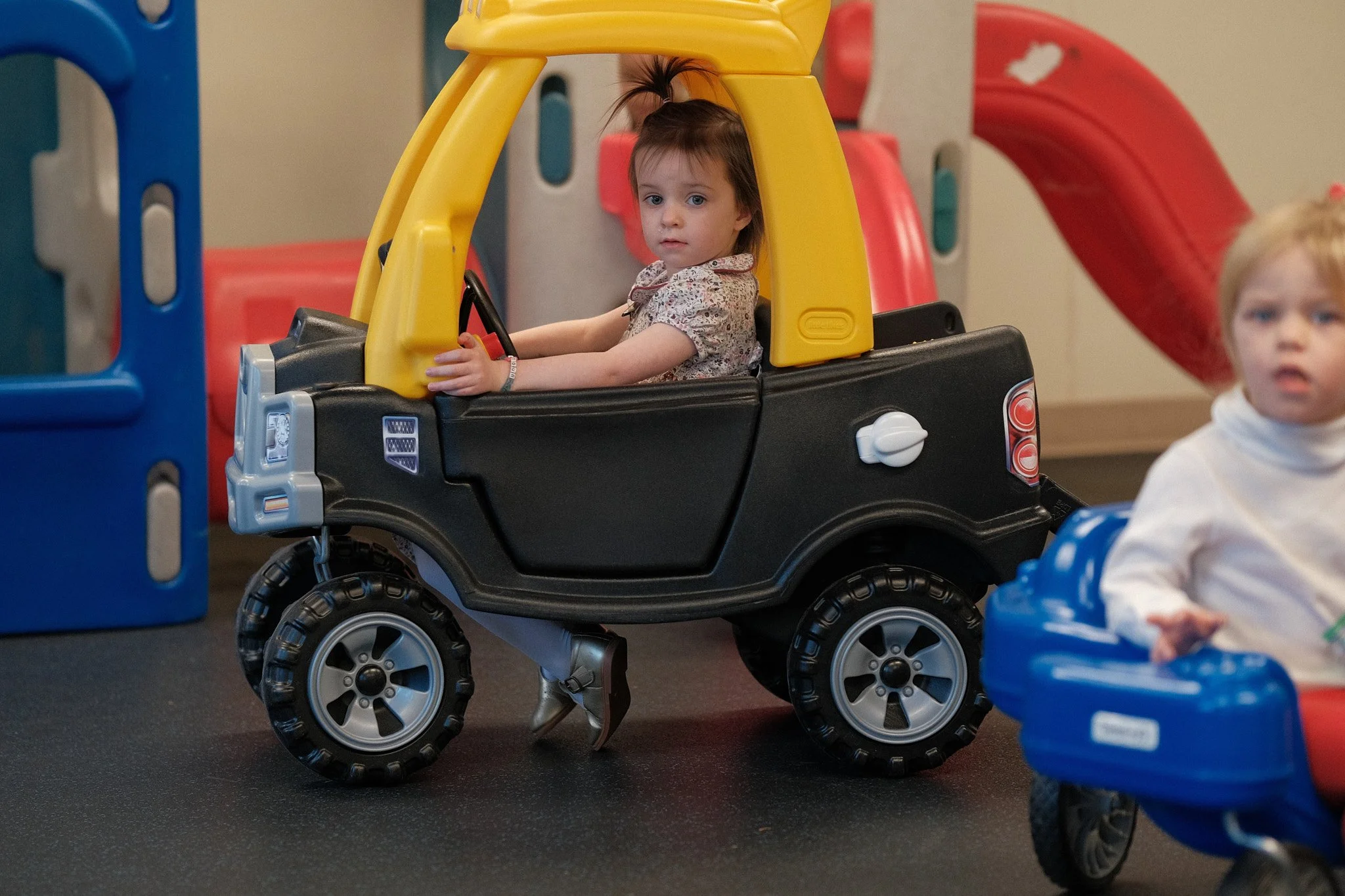 Two young children playing at an indoor playground, with one girl sitting in a toy vehicle resembling a black and yellow truck, and another girl in a blue and red stroller-like toy next to her.