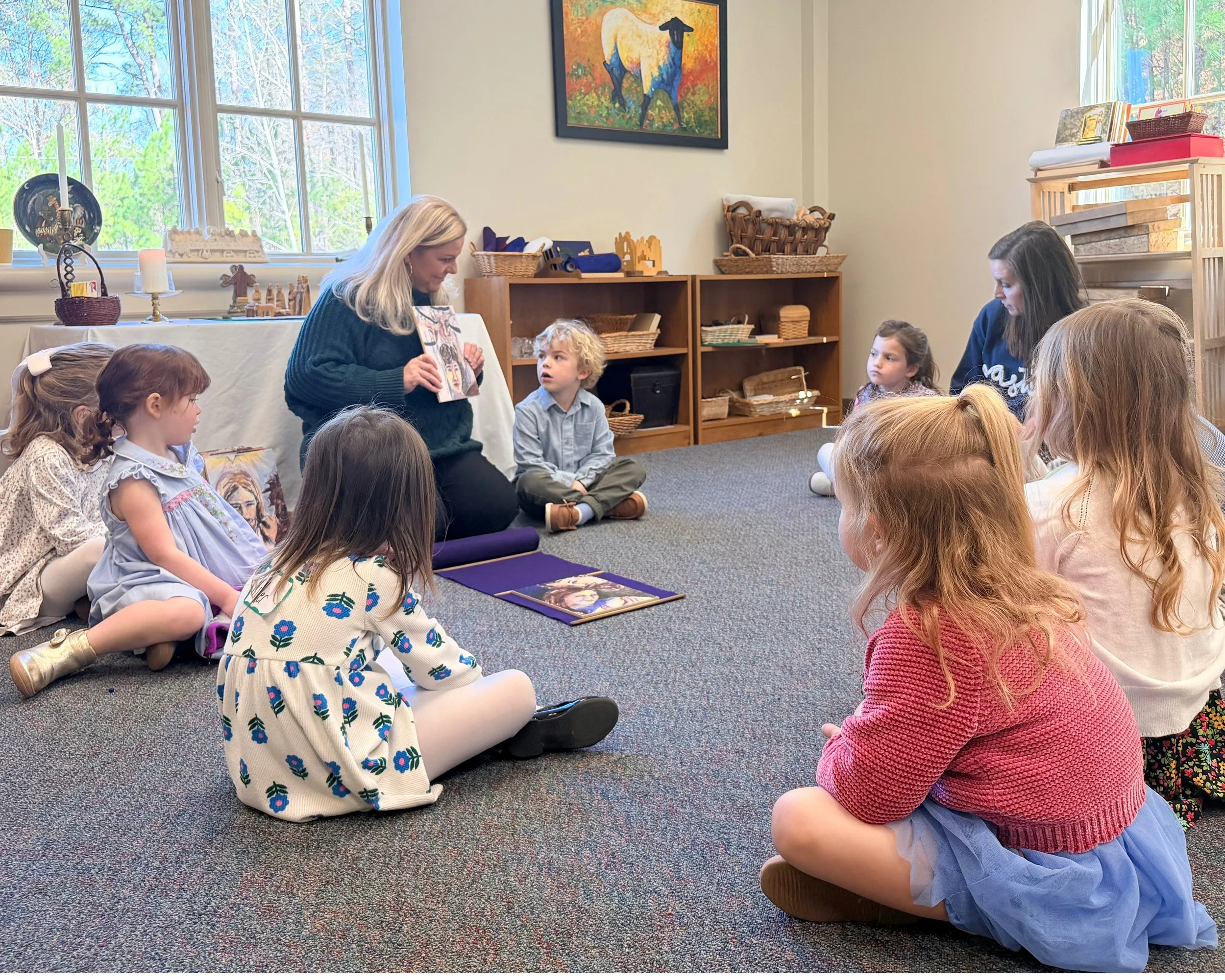 A woman reading a picture book to a group of young children seated on a carpeted floor in a cozy room with large windows, shelves, and colorful artwork on the walls.