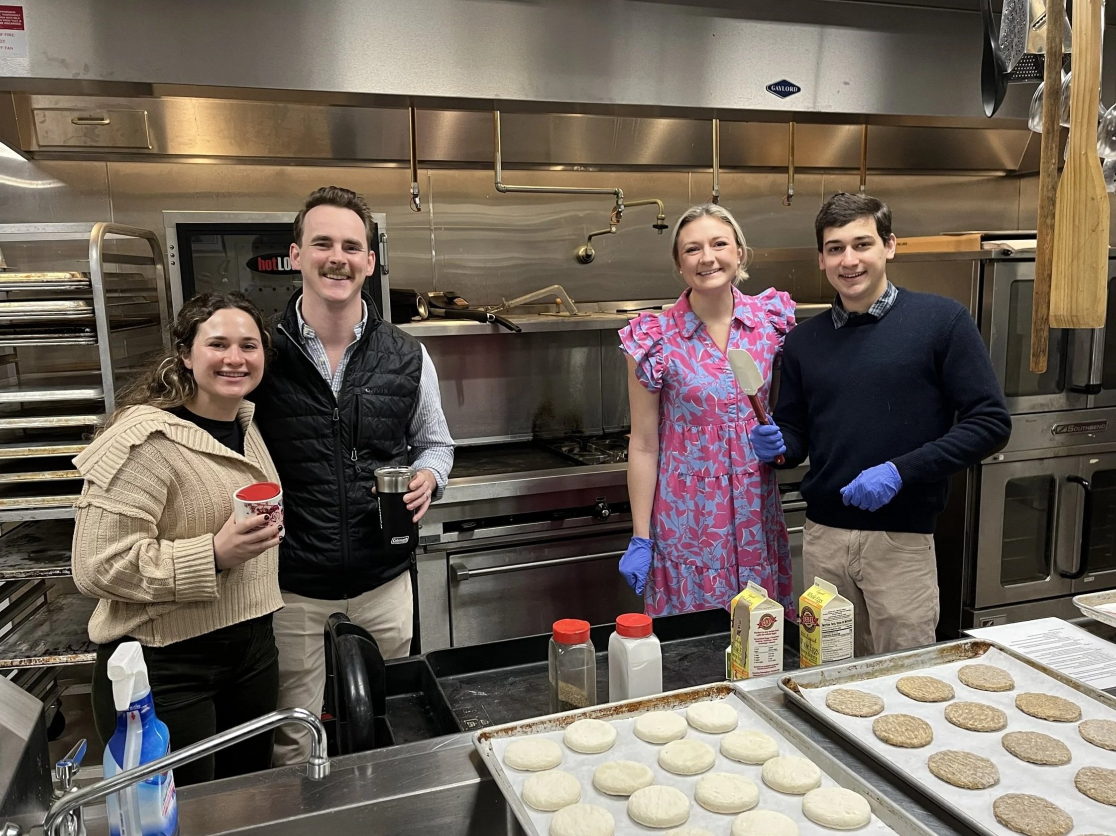 Four people in a kitchen, smiling, standing behind baking trays with partially baked cookies. Two are holding cups, and one is holding a spatula. They appear to be preparing baked goods.