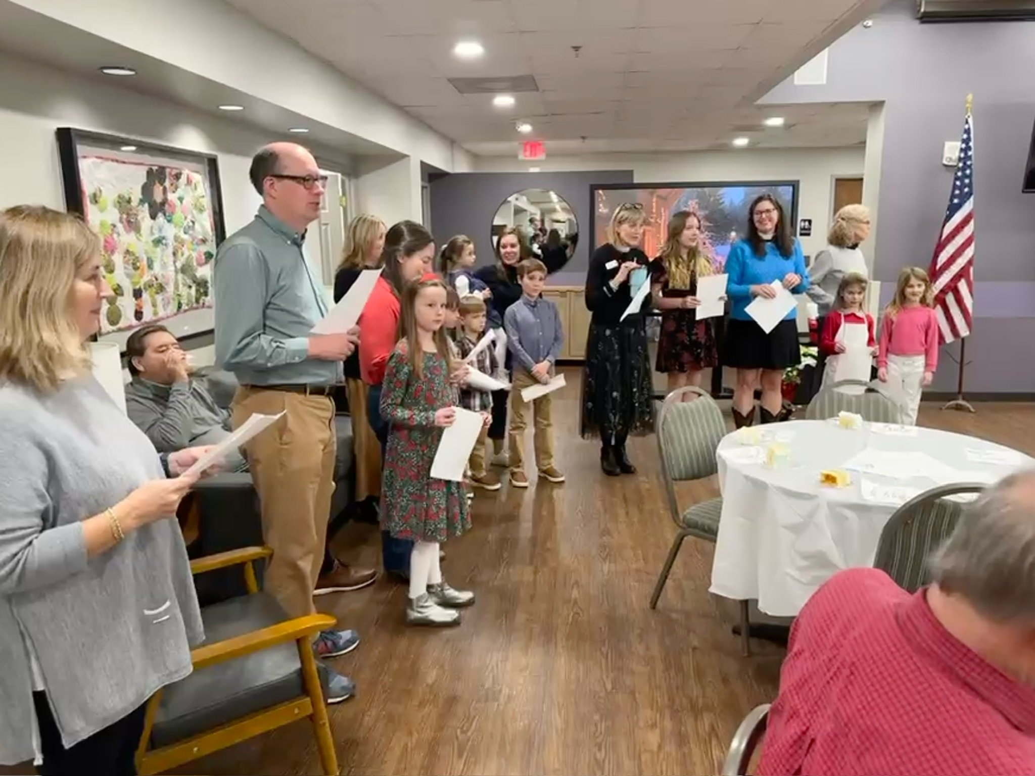Group of children and adults standing in a room, some holding papers, participating in a ceremony or presentation in a decorated event space with round tables, American flag, and artwork on the walls.