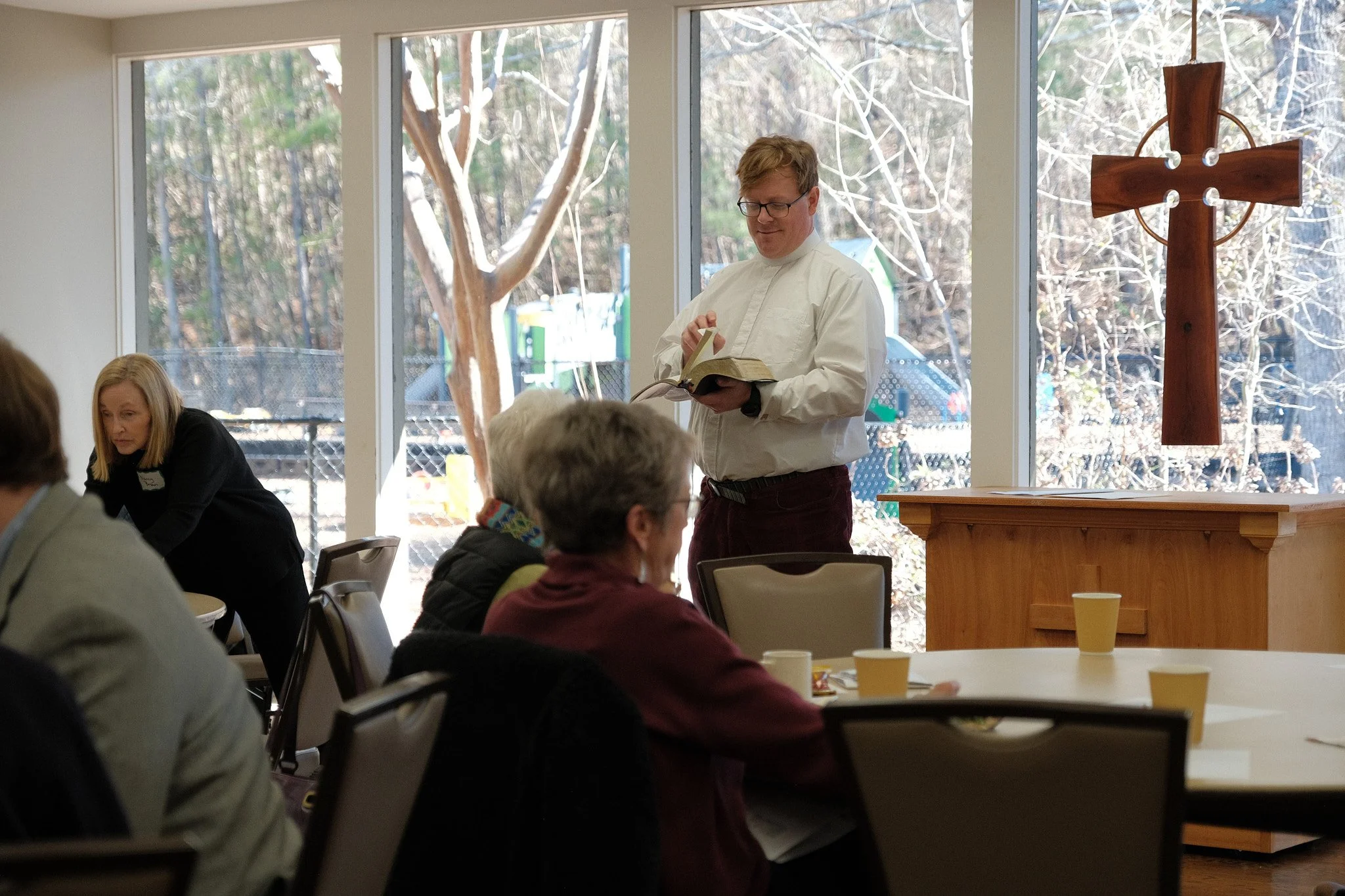 A man reading a book at a gathering in a room with large windows and a cross hanging on the wall. Several people are seated at tables with cups.