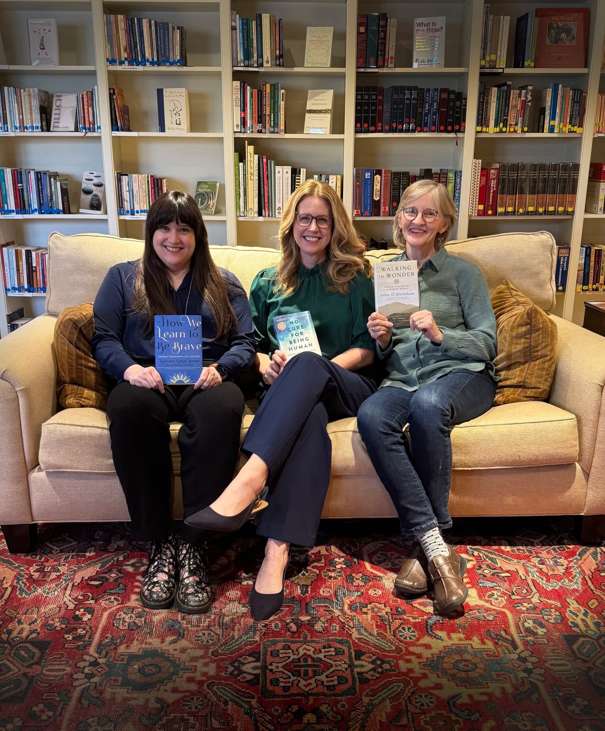 Three women sitting on a beige sofa in a library, each holding a book and smiling at the camera. The library has multiple bookshelves filled with books in the background.
