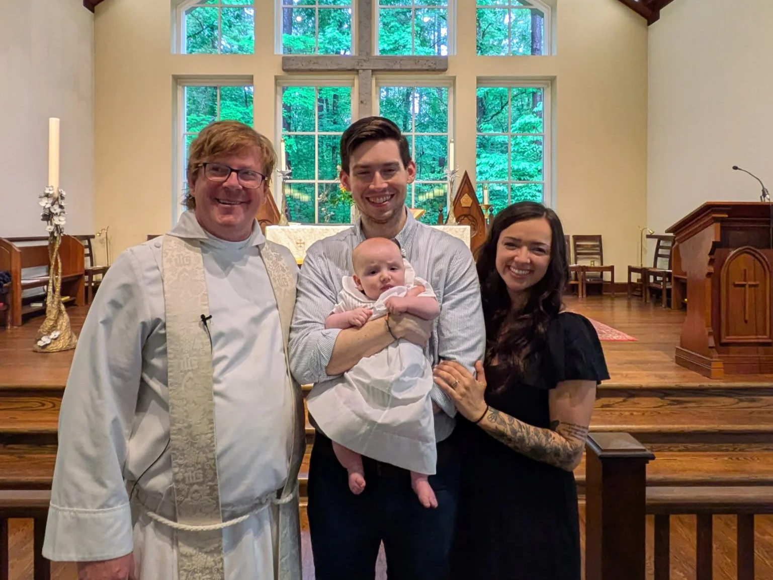 A family standing together inside a church, posing for a photo. The family includes a priest, a man holding a baby, and a woman. The church's large window behind them shows green trees outside.