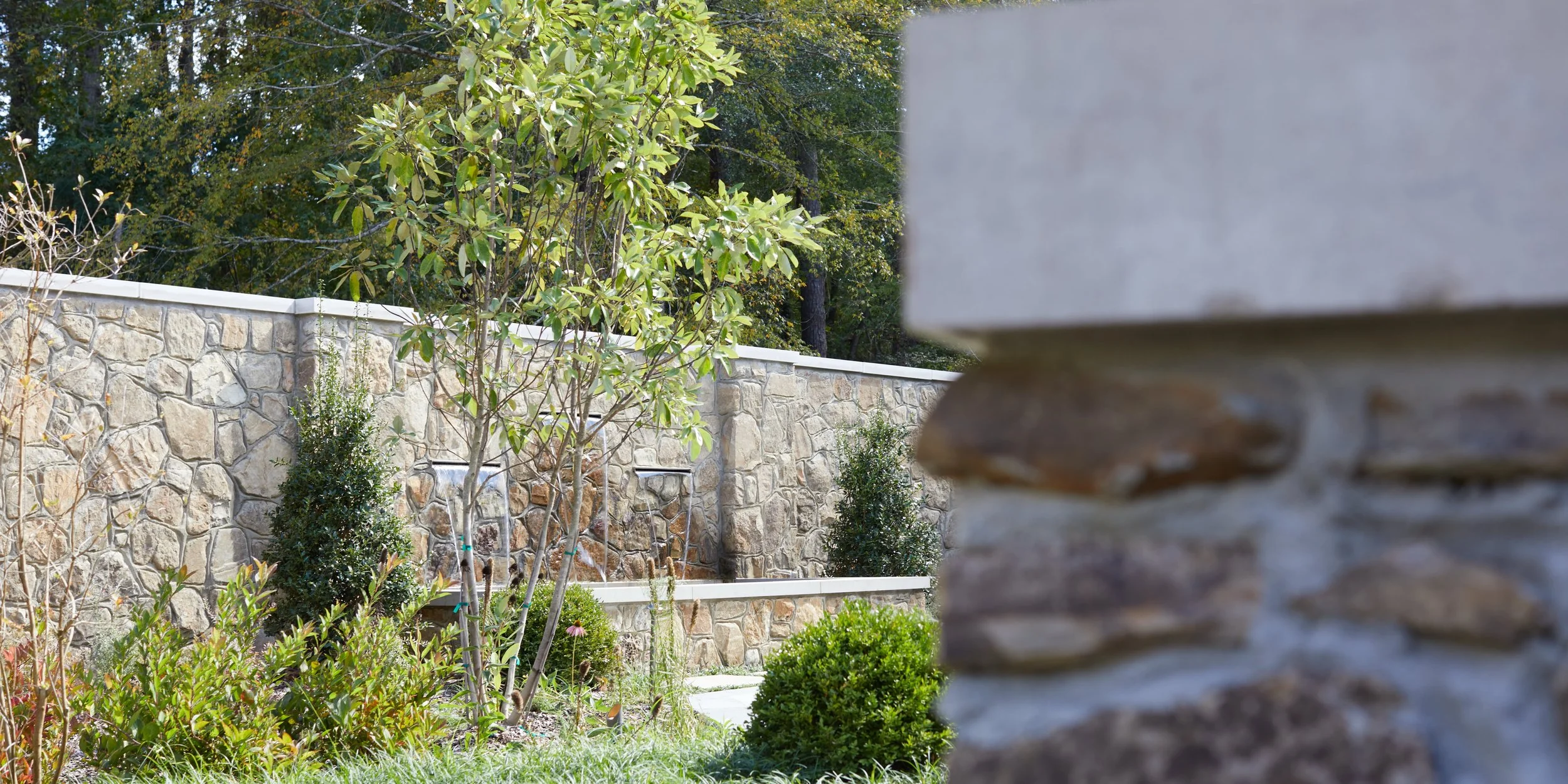 A stone wall with a white trim in a garden, with green trees and bushes in the background and some plants and small trees in the foreground.