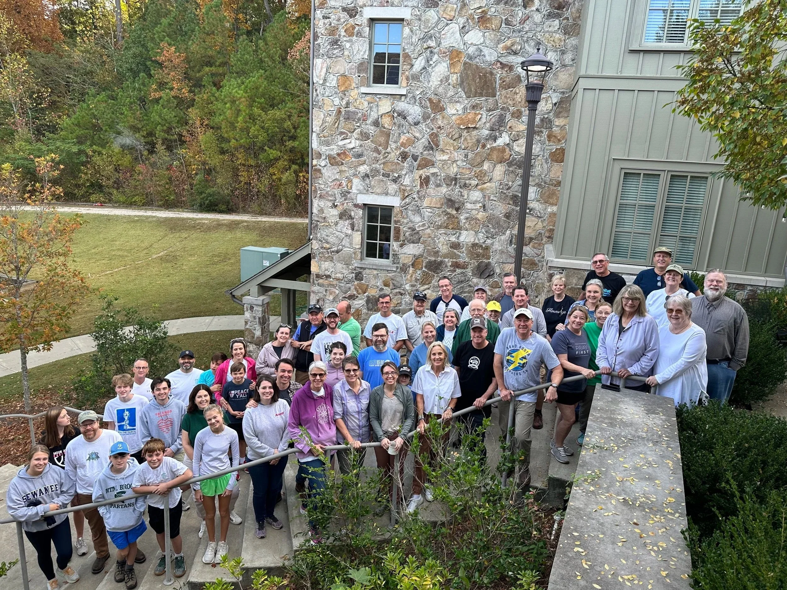 A large group of people gathered outdoors on a concrete staircase and walkway, posing for a group photo in front of a stone building with large windows and a lamp post, with trees and a grassy area in the background.