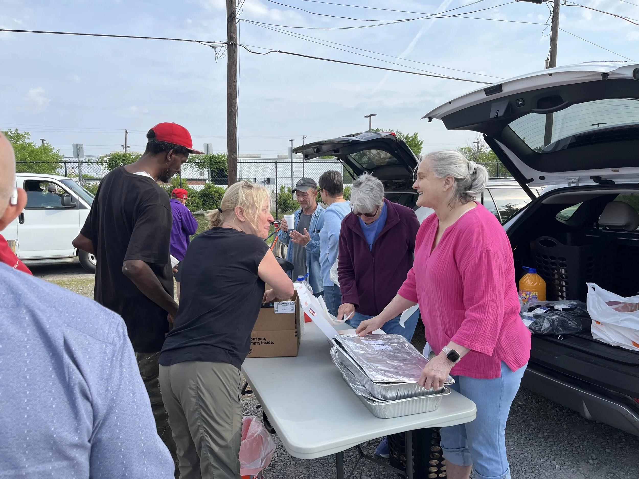 People gathered around a table outdoors near parked cars, serving and receiving food at a community meal or event.
