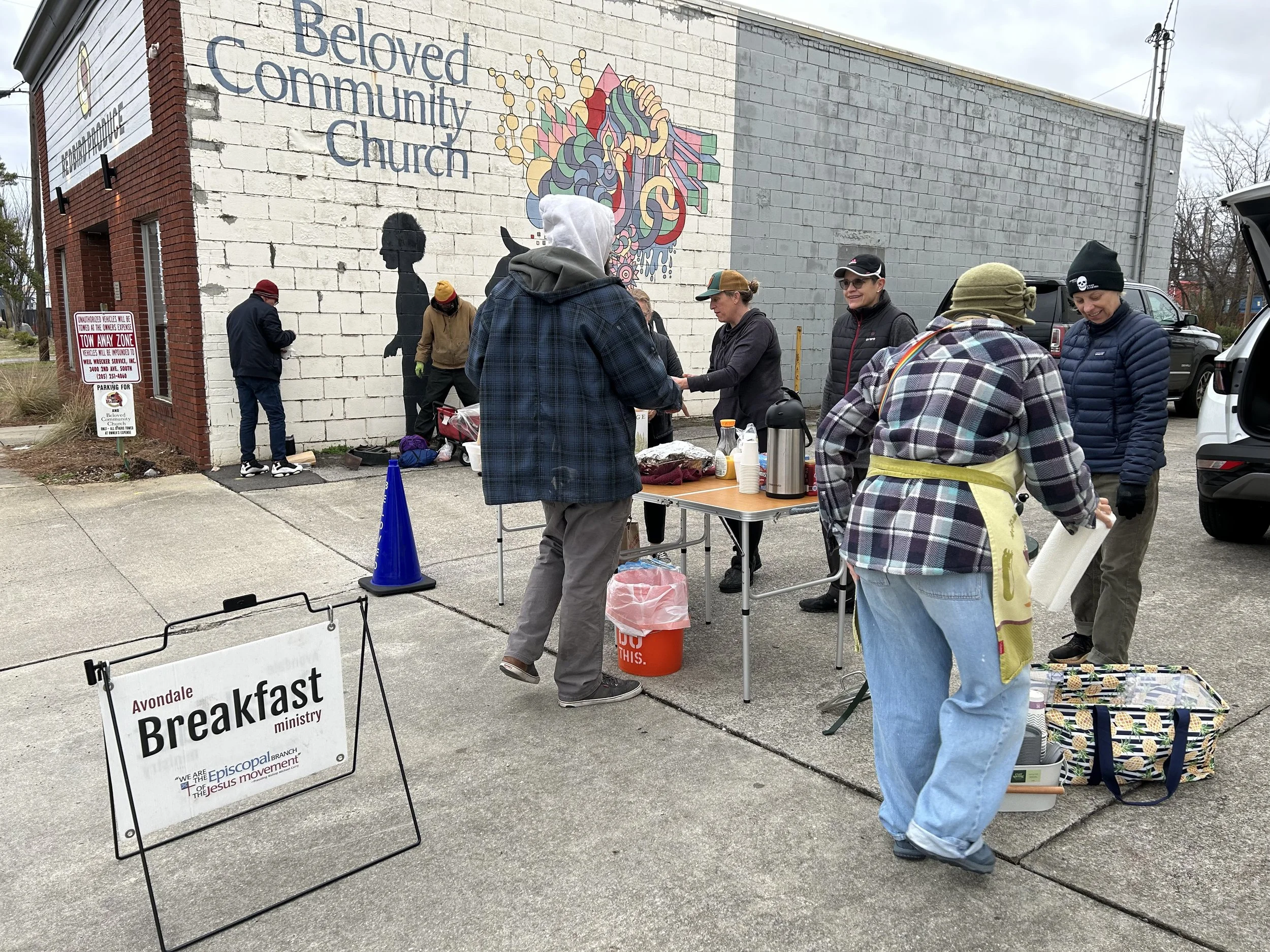 People gathered outside Beloved Community Church for a breakfast ministry, with tables set up for serving food, and a sign indicating 'Avondale Breakfast Ministry.'