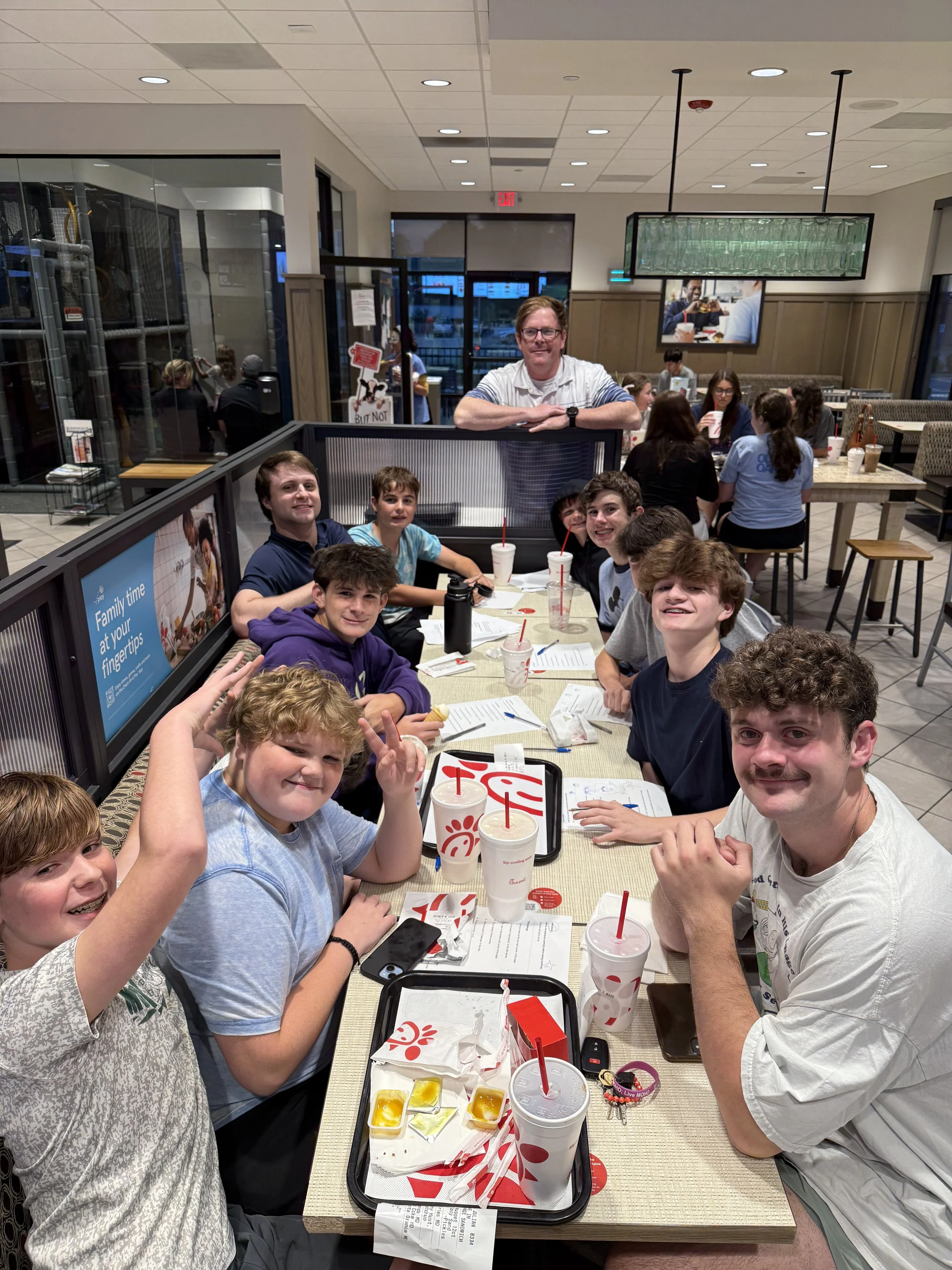 Group of young boys and two adults sitting at a table in a Chick-fil-A restaurant, with food and drinks on the table, smiling and looking at the camera.