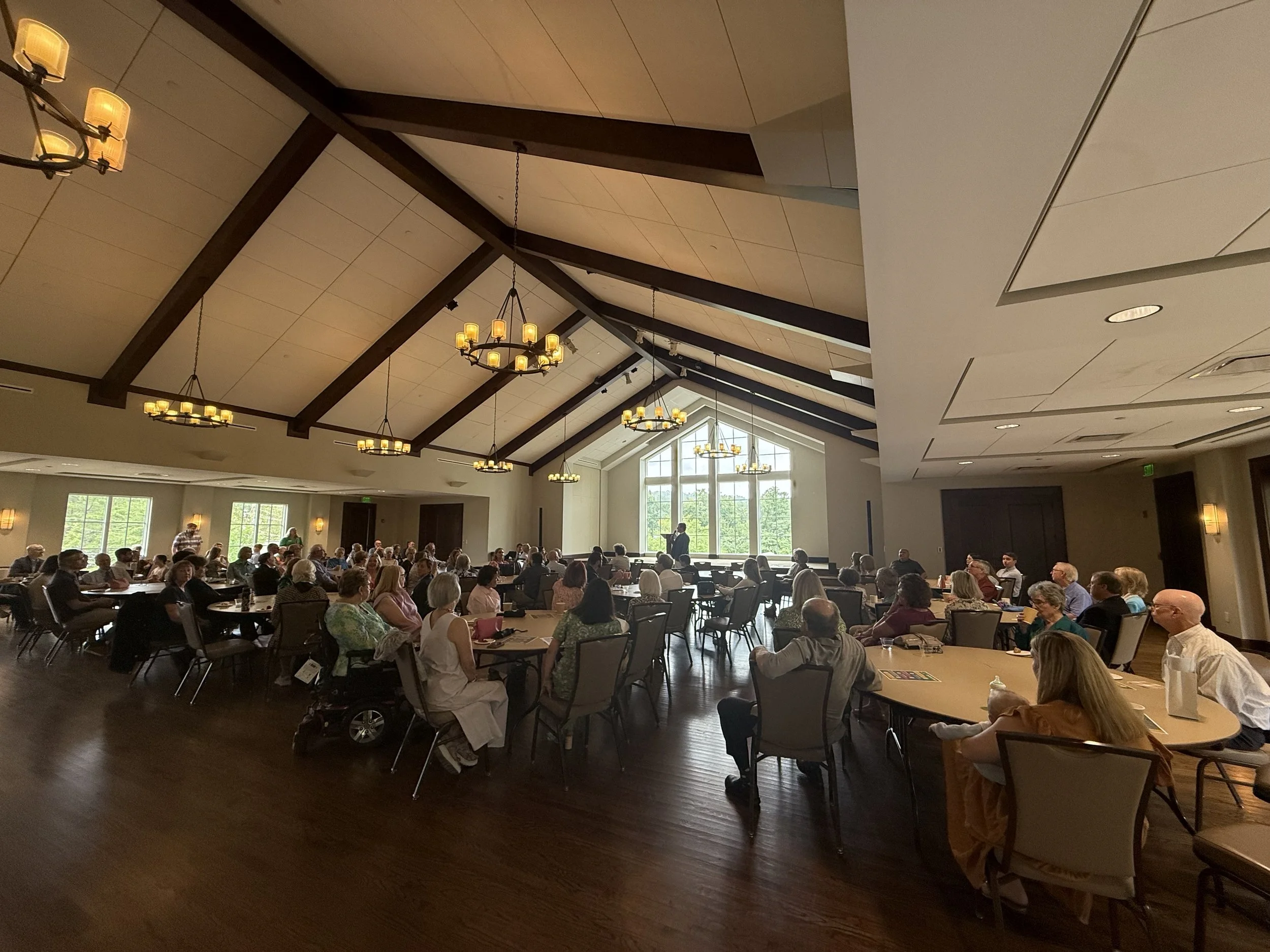 Meeting or conference taking place in a large, well-lit room with high, vaulted ceilings and chandeliers, where many people are seated at round tables listening to a speaker at the front near large windows.