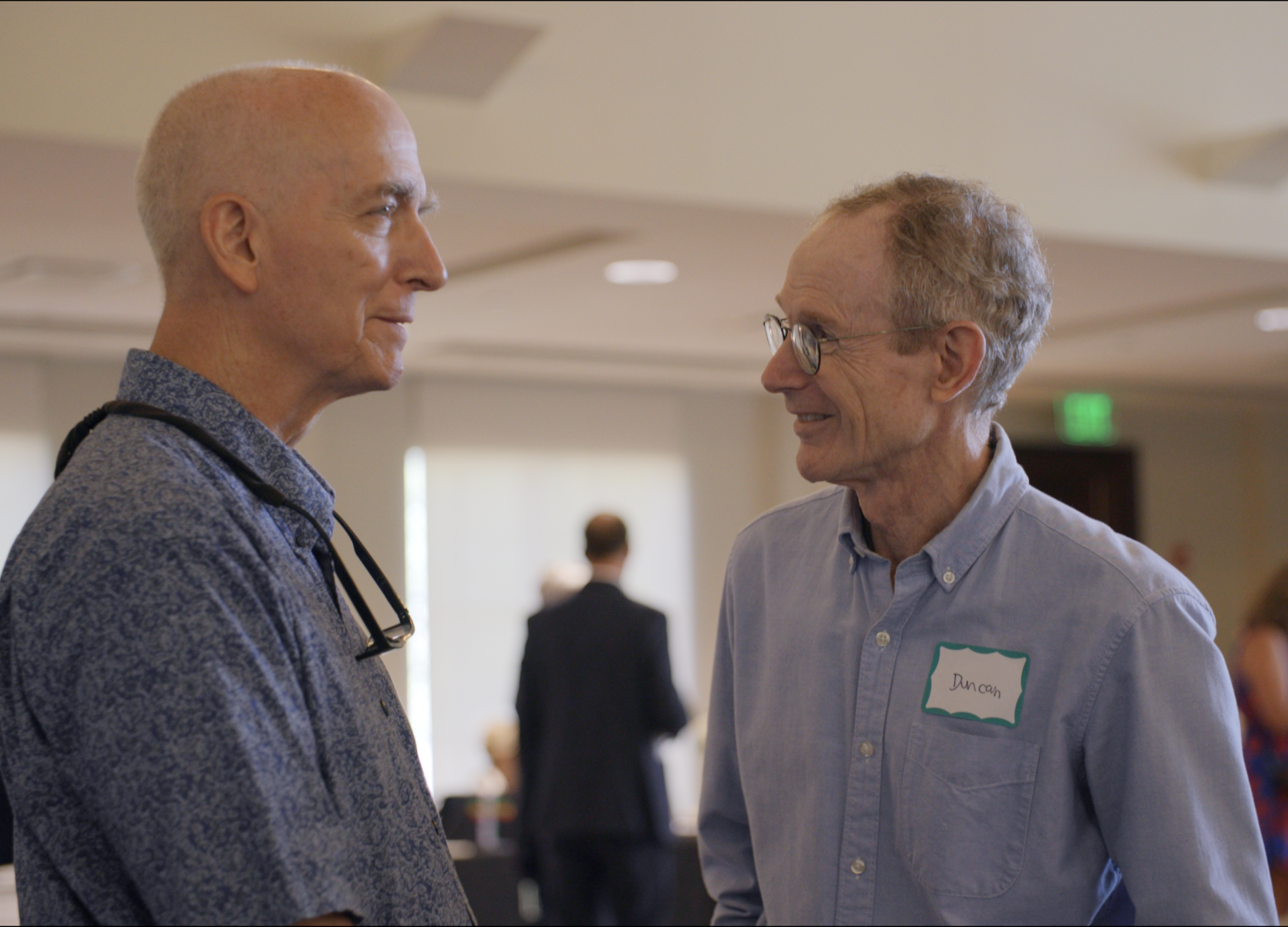 Two elderly men are smiling and talking with each other at a social event in a brightly lit room. One man is wearing a patterned shirt with glasses hanging around his neck, and the other man is wearing a light blue shirt with a name tag that reads "Duncan."