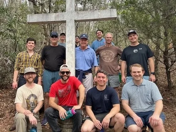 Group of ten men outdoors in a wooded area, standing and sitting around a large T-shaped cross.