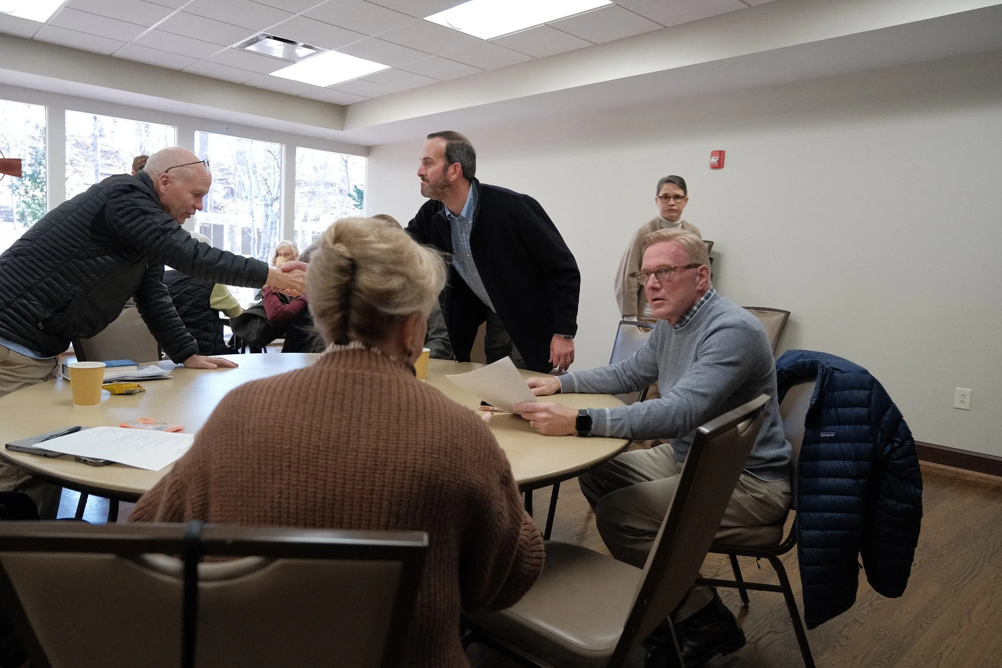 People sitting around a table in a meeting room with large windows in the background, some engaging in conversation and shaking hands.