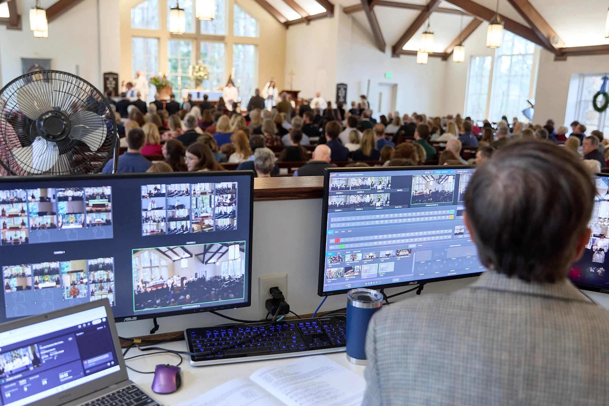A person operating video editing equipment at the back of a church during a service or event, with a large congregation seated inside.