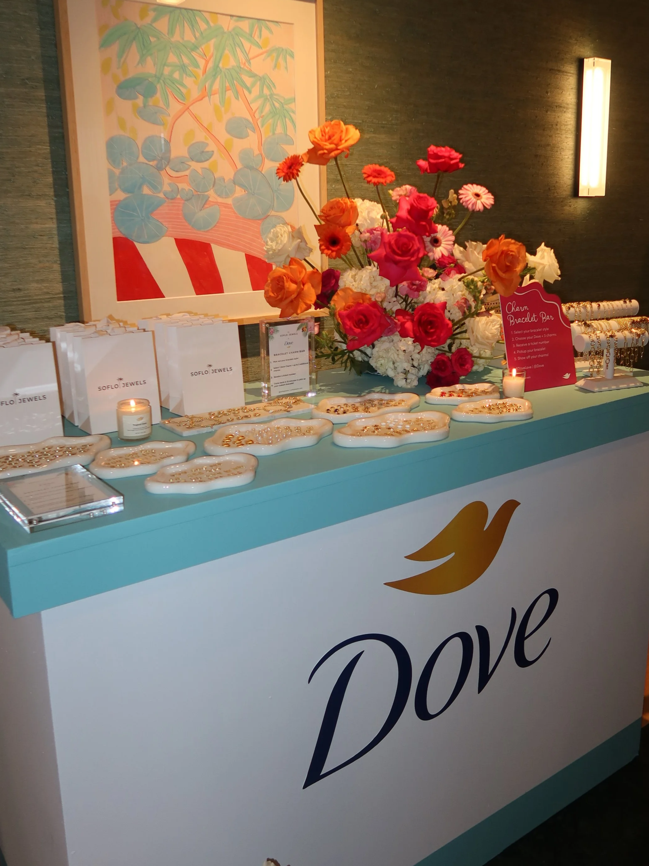 A display table with Dove branding, decorated with a large colorful flower arrangement, jewelry, and promotional materials, likely set up for an event or promotional activity. on-site charm bar custom charm bracelets miami events 
