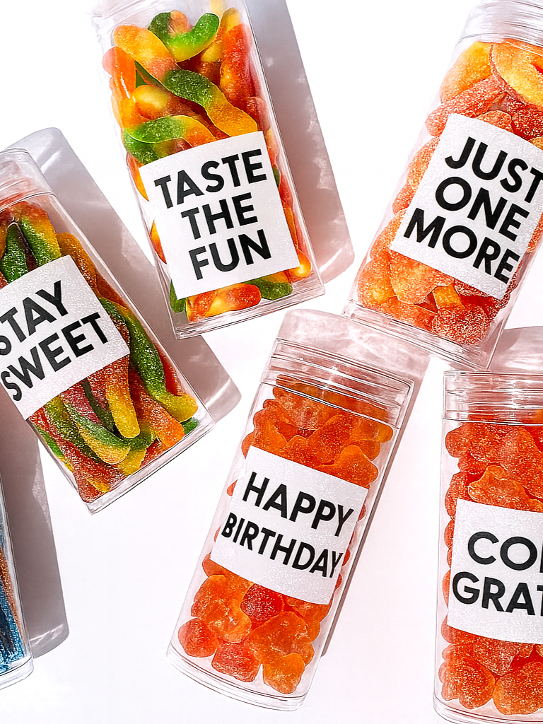 Clear plastic containers filled with colorful gummy candies, each with a label containing celebratory or fun messages like "Taste the Fun," "Just One More," "Stay Sweet," and "Happy Birthday." custom branded candy gifting corporate event activation