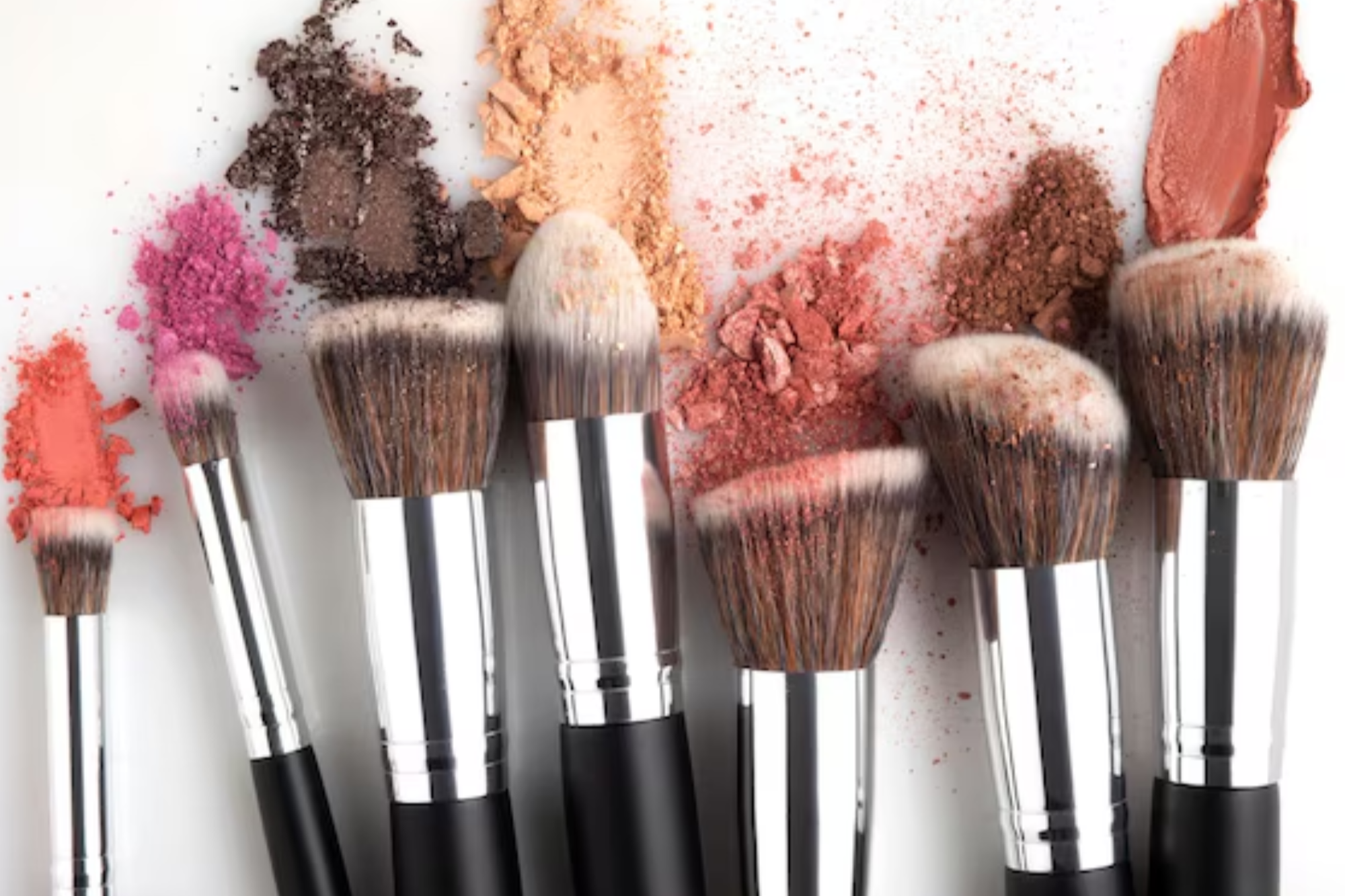 Makeup brushes with colorful powder eyeshadows and blushes scattered on white background.