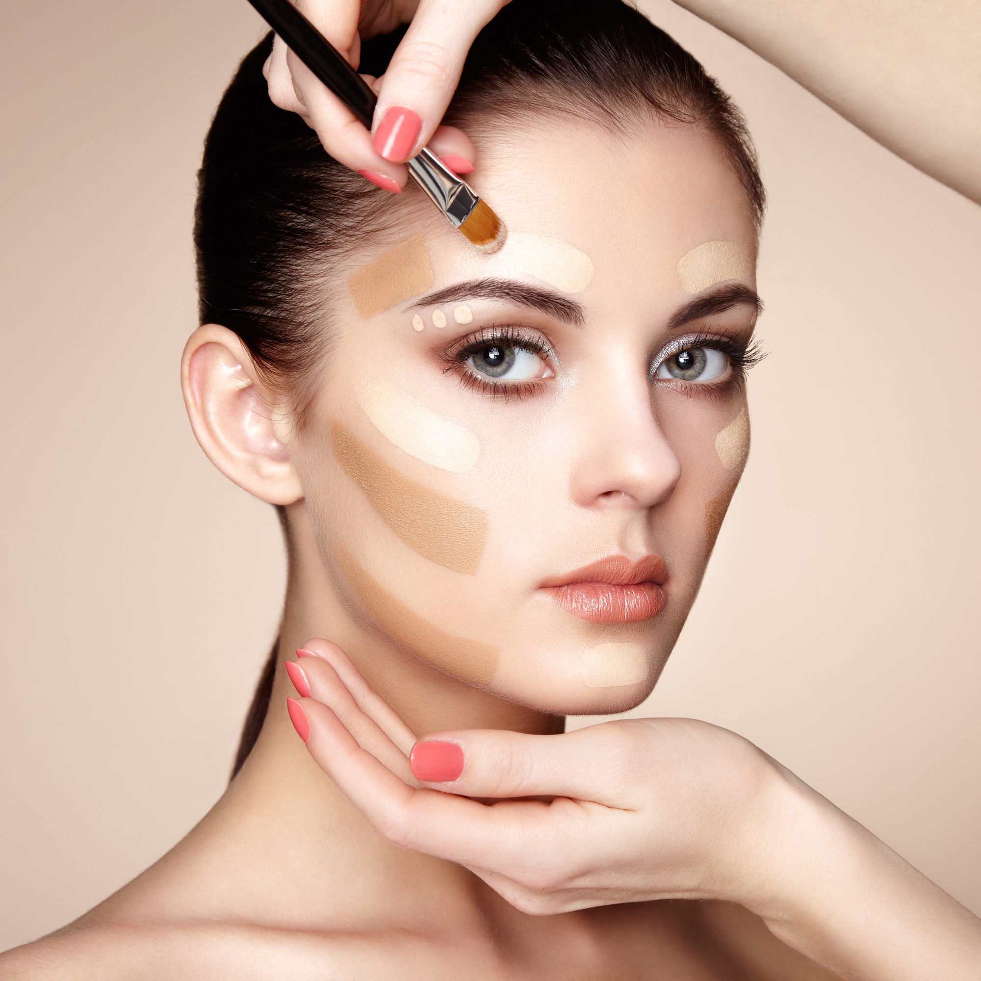 A woman with blue eyes and dark hair is having various shades of foundation applied to her face and forehead by a makeup artist, with the artist's hand visible applying makeup with a brush.