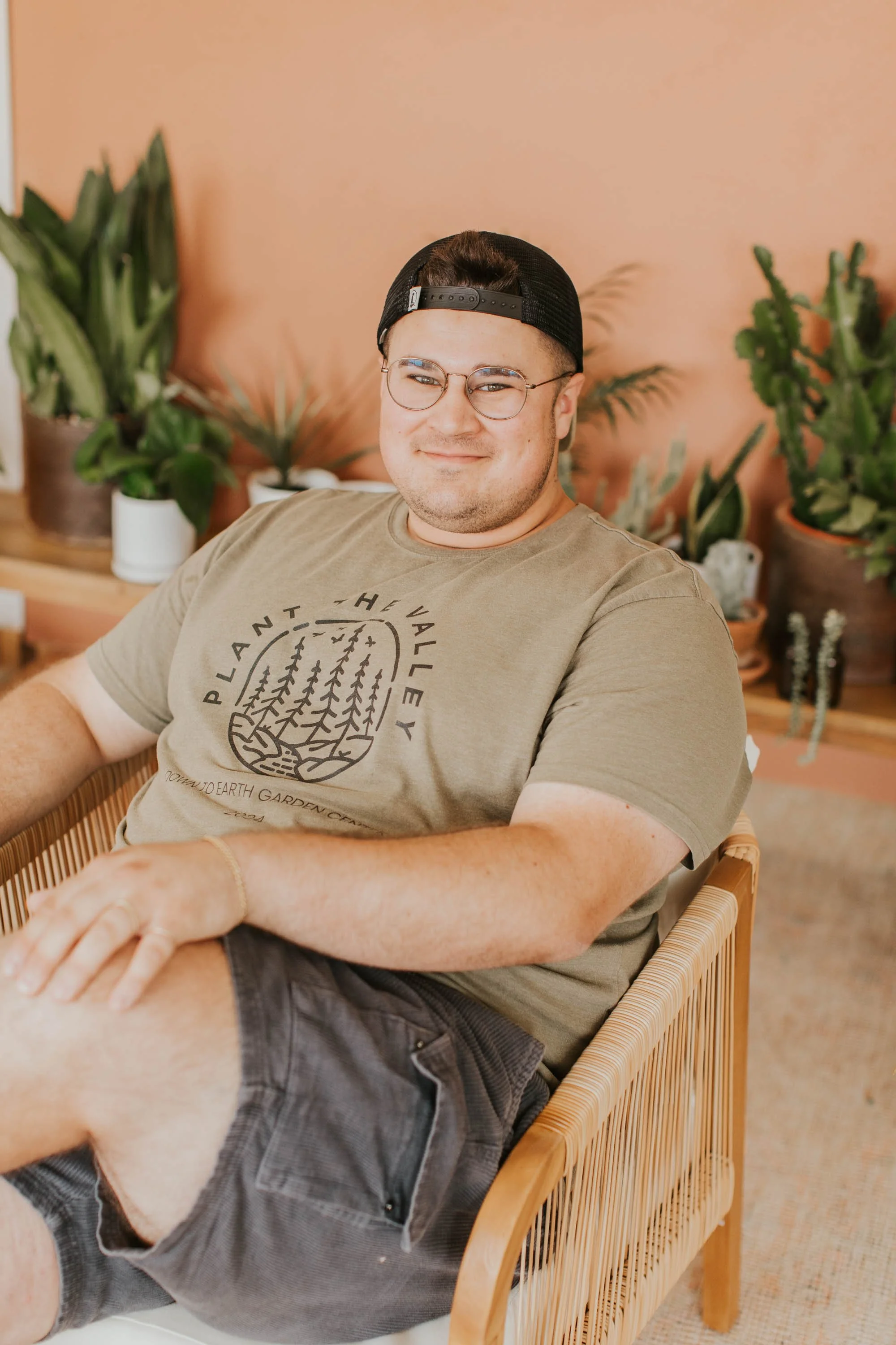 A young man wearing glasses, a black cap backwards, a beige t-shirt with a forest design, and dark shorts, sitting in a wooden chair surrounded by potted plants against a peach-colored wall, smiling at the camera.