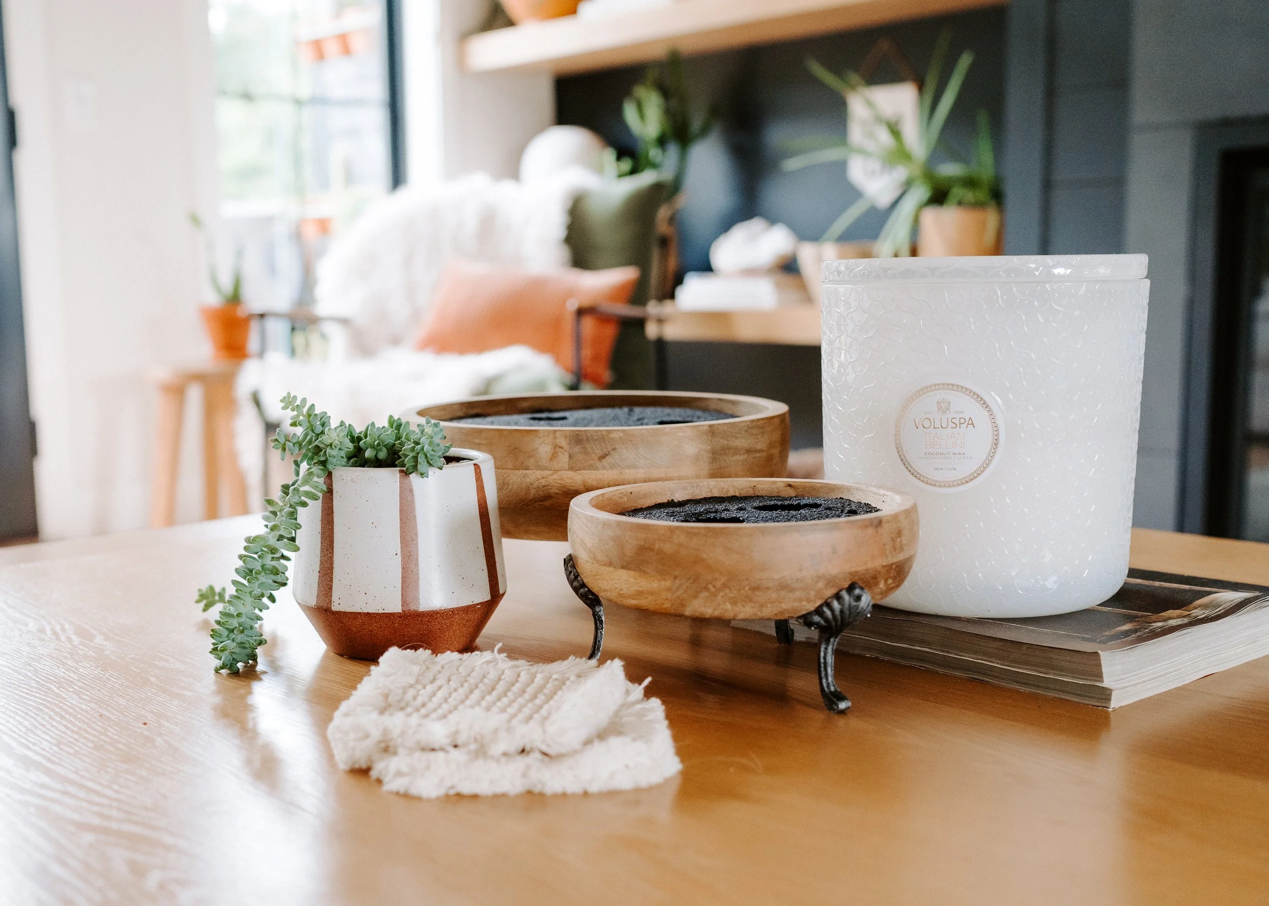 Close-up of a wooden table with small potted succulent plants, candles, and a dish, with a blurred background of a cozy living space with cushions, plants, and natural light from windows.