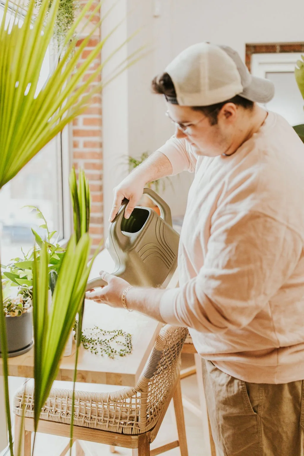 A man is watering plants in a sunlit room with large windows, a brick wall, and various green potted plants.