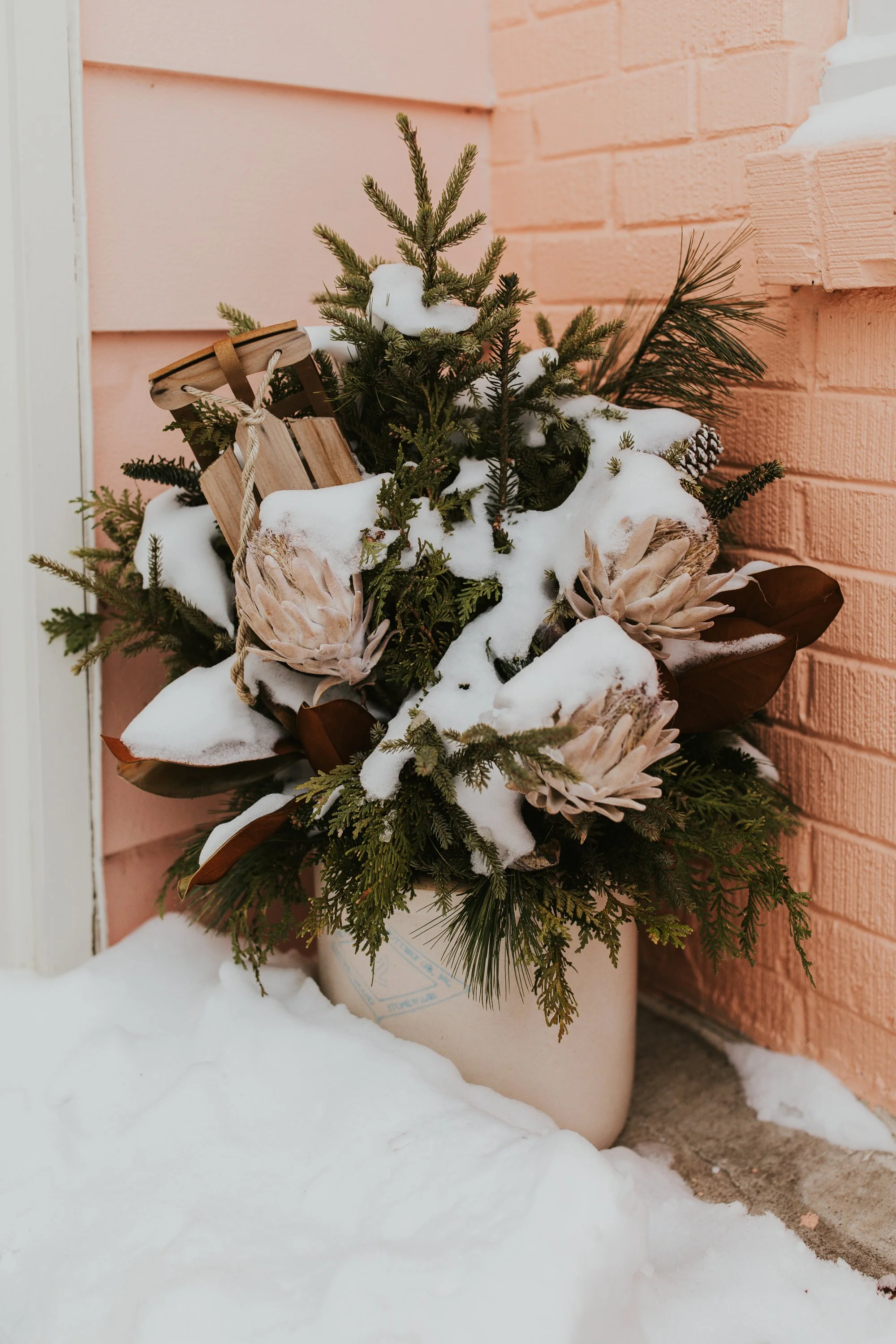 A small Christmas tree with snow on its branches, decorated with a wooden sled and large pinecones, placed outdoors against a pink brick wall and surrounded by snow.