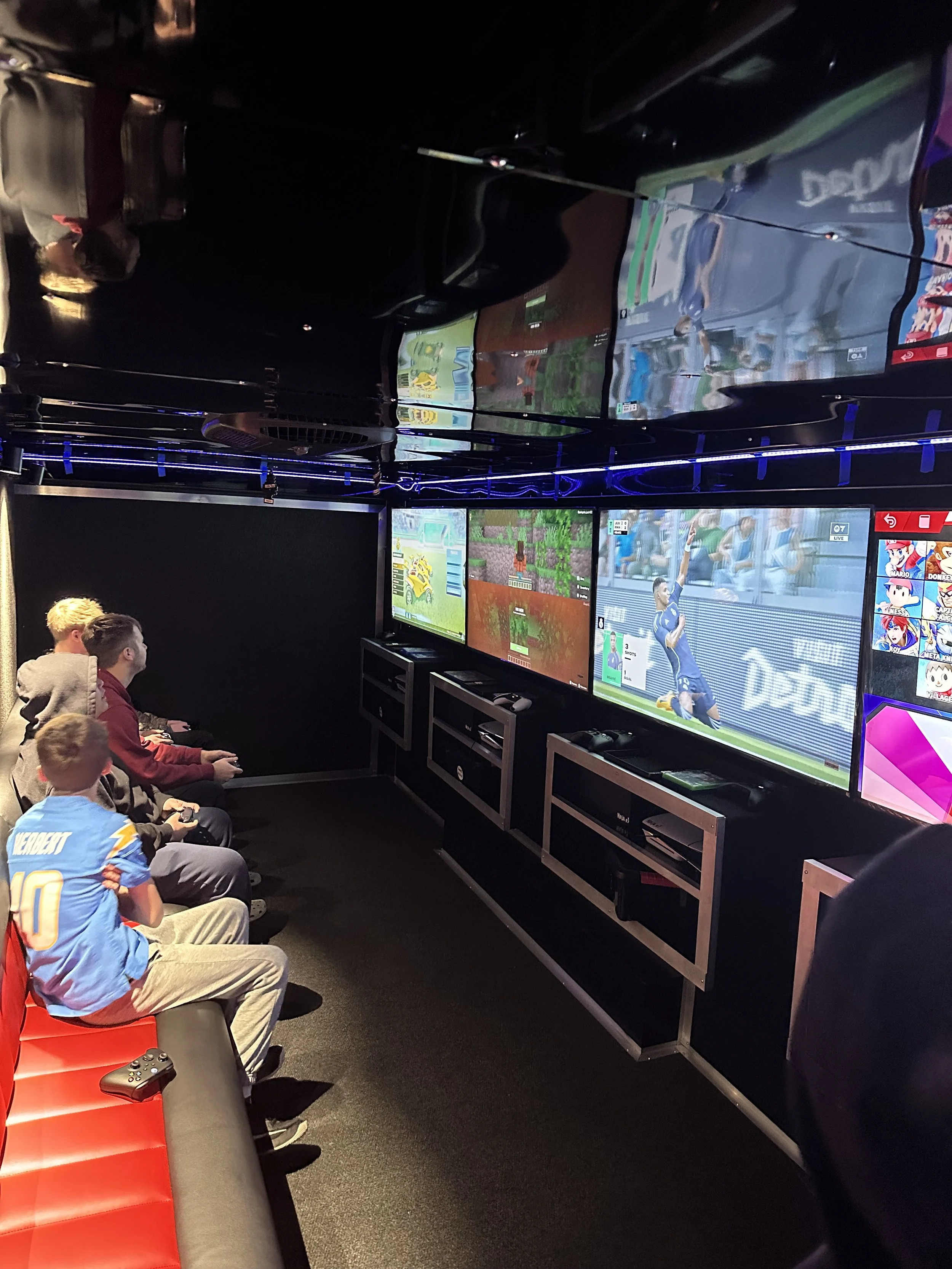 Group of children and a man playing video games on multiple screens in a gaming trailer or gaming truck rental with black walls and ambient purple lighting a church Christmas party.